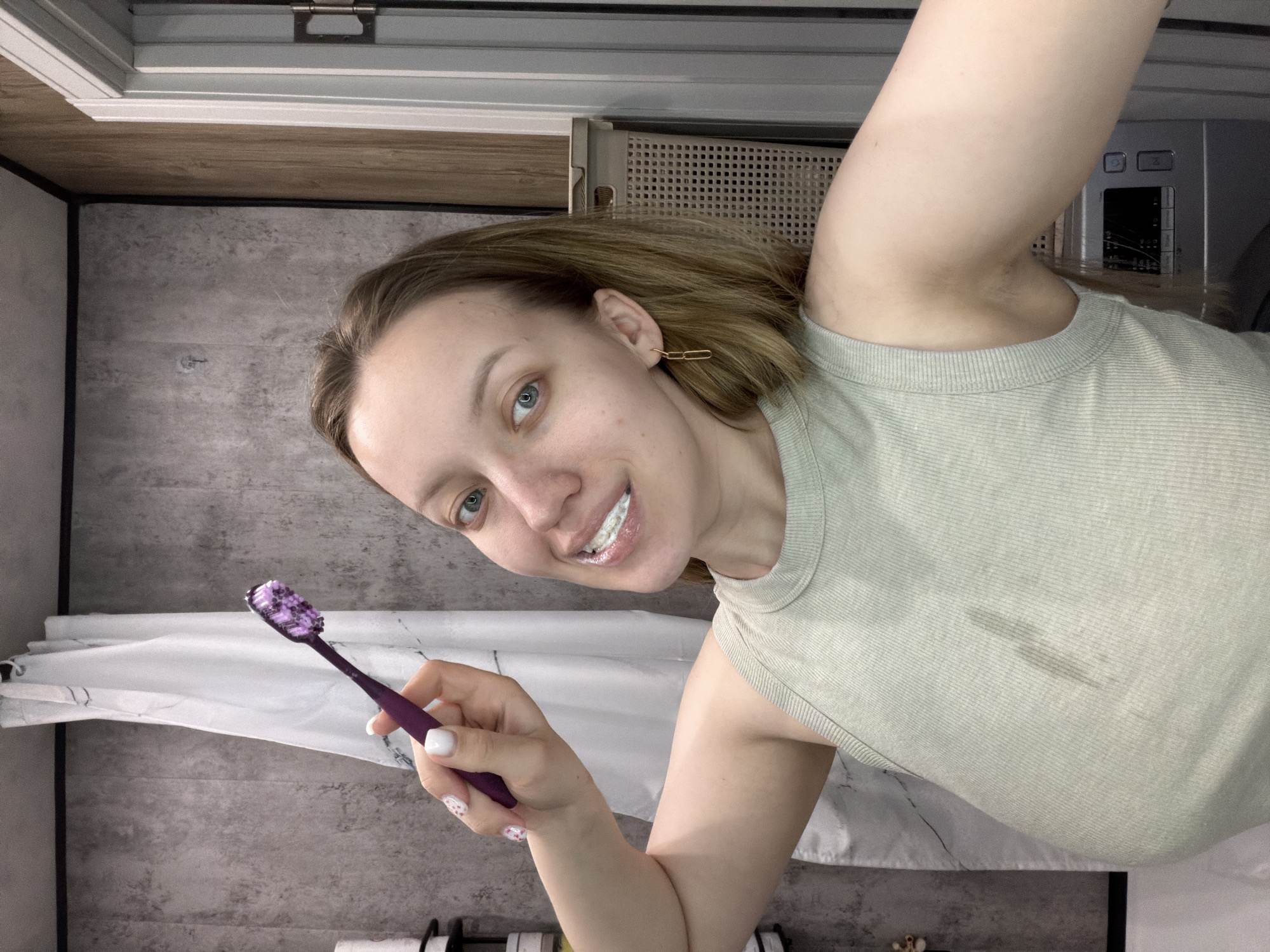 A female subject is depicted in what appears to be a bathroom located in Reshetnikovo, Russia. The subject has light skin, blue eyes, and light brown, shoulder-length hair. She is smiling, with white toothpaste foam visible around her teeth. She wears a light green, ribbed tank top, which shows a dark stain on the front chest area. A gold-colored chain-link earring is present on her right ear. Her left hand is raised, holding a purple toothbrush with purple toothpaste on its bristles. The visible fingernails on her left hand are painted white, with her ring finger displaying a white base with small colorful dots.
The background consists of a white shower curtain with a grey marbled pattern, hanging from a rod against a dark grey textured wall paneling. To the far left, a black wall shelf with white containers is partially visible. To the right, a light brown, woven plastic hamper or basket is present, with a silver-colored appliance resembling a washing machine partially visible below it, behind the subject's right arm. A white door frame with a visible hinge and adjacent wooden wall paneling occupies the right side of the image. A small metallic hook is affixed to the grey wall paneling in the upper left background.