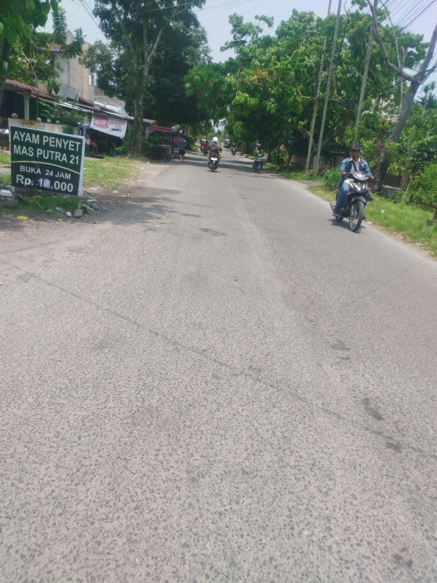 A person on a motorcycle is seen riding on a road in front of a sign that reads "AYAM PENYET MAS PUTRA 21 BUKA 24 JAM Rp. 10.000" which translates to "Chicken Smash Mas Putra 21 Open 24 Hours Rp. 10,000". The road is paved and has some markings. On the side of the road, there are trees and some buildings. The sky is clear and it appears to be daytime. The image is taken from a low angle, looking up at the motorcycle and the road.