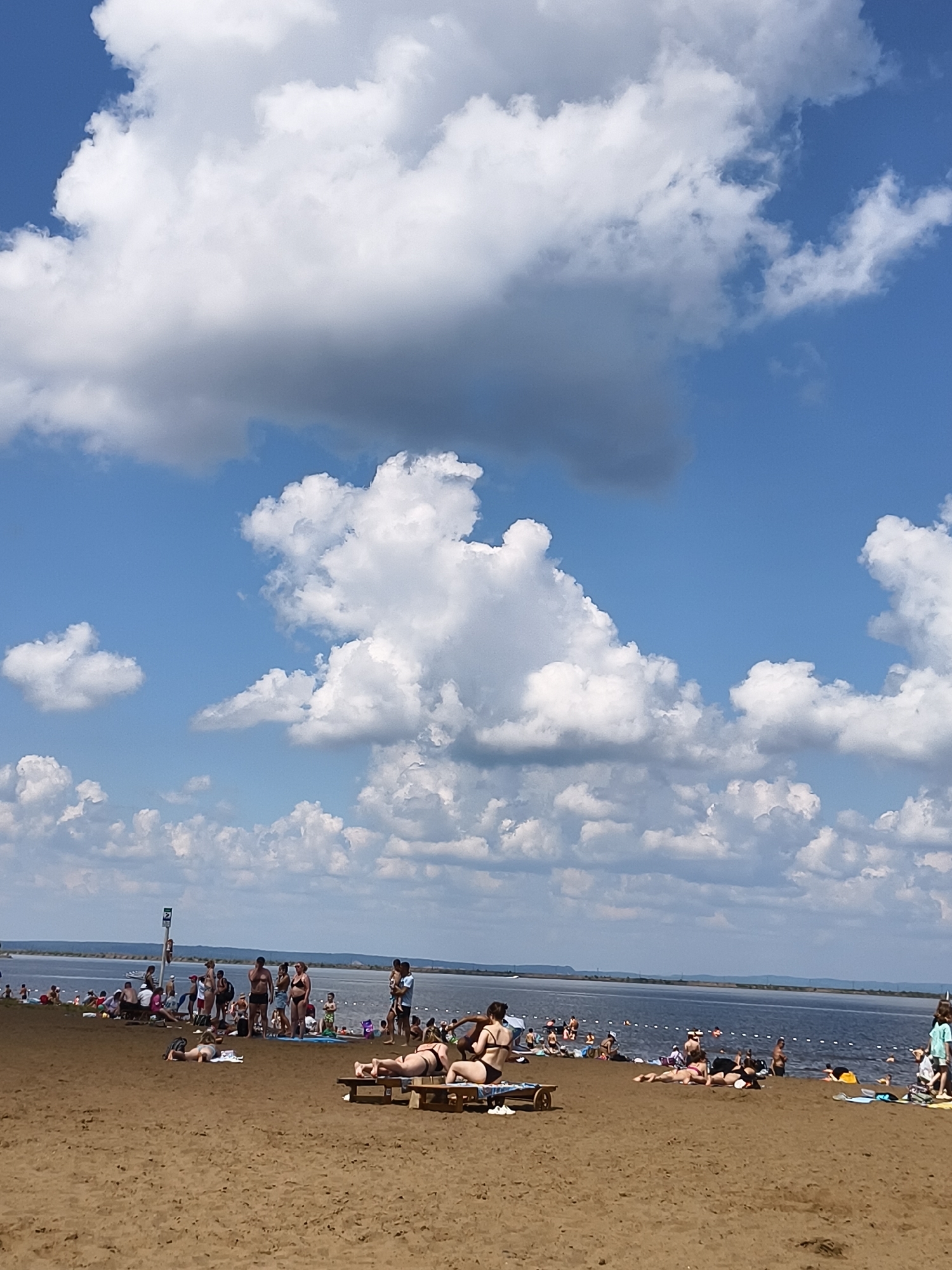 The image shows a crowded beach on a sunny day with scattered clouds. People are sunbathing on the sand and in the water. Some individuals are lying on towels or beach chairs, while others are standing or walking. The water is blue and appears calm, with a thin line of buoys in the distance. The beach itself is sandy and stretches towards the water. In the background, there are more people gathered near the water's edge, and a distant shoreline is visible. The sky is a bright blue with large, fluffy white clouds. There is no discernible city or specific landmark that would indicate a location.