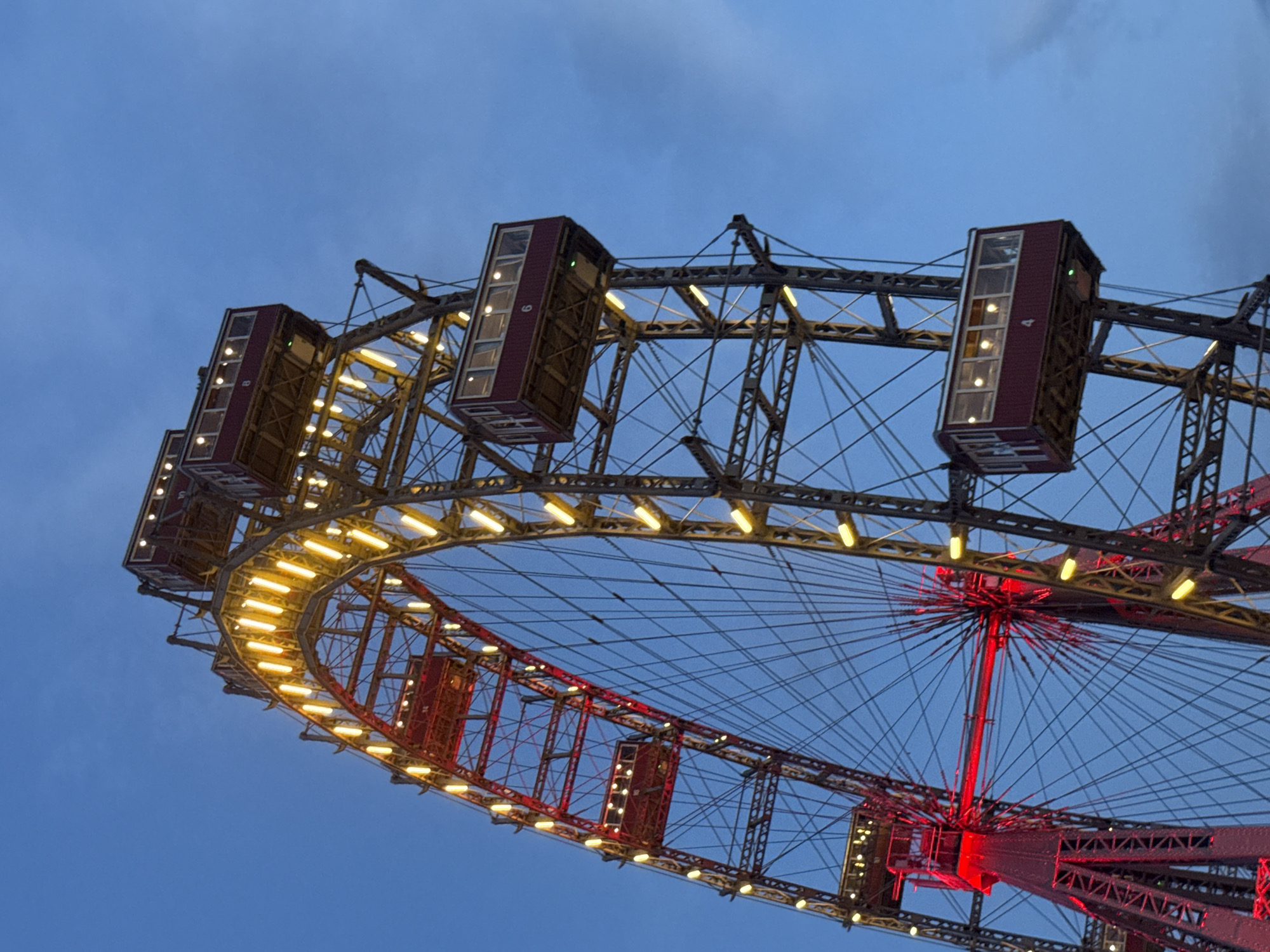 This is a low-angle, exterior, medium shot of the Wiener Riesenrad, a Ferris wheel located in the Prater amusement park in Vienna, Austria. The sky is a deep blue, transitioning to lighter shades as it moves towards the horizon, with a few wispy clouds. The Ferris wheel, a prominent landmark, is illuminated with a combination of red and yellow lights. The red lights are concentrated on the lower structural elements and central hub, while yellow lights line the outer rim and highlight the individual cabins. One cabin, numbered "6," is clearly visible in the upper right portion of the frame. Another cabin, numbered "4," is visible in the lower right. The intricate metal latticework of the wheel's structure is visible, along with the numerous cables that support the cabins. The scene is captured at dusk or twilight, as indicated by the dim ambient light and the illuminated structure. There are no people visible in the image.