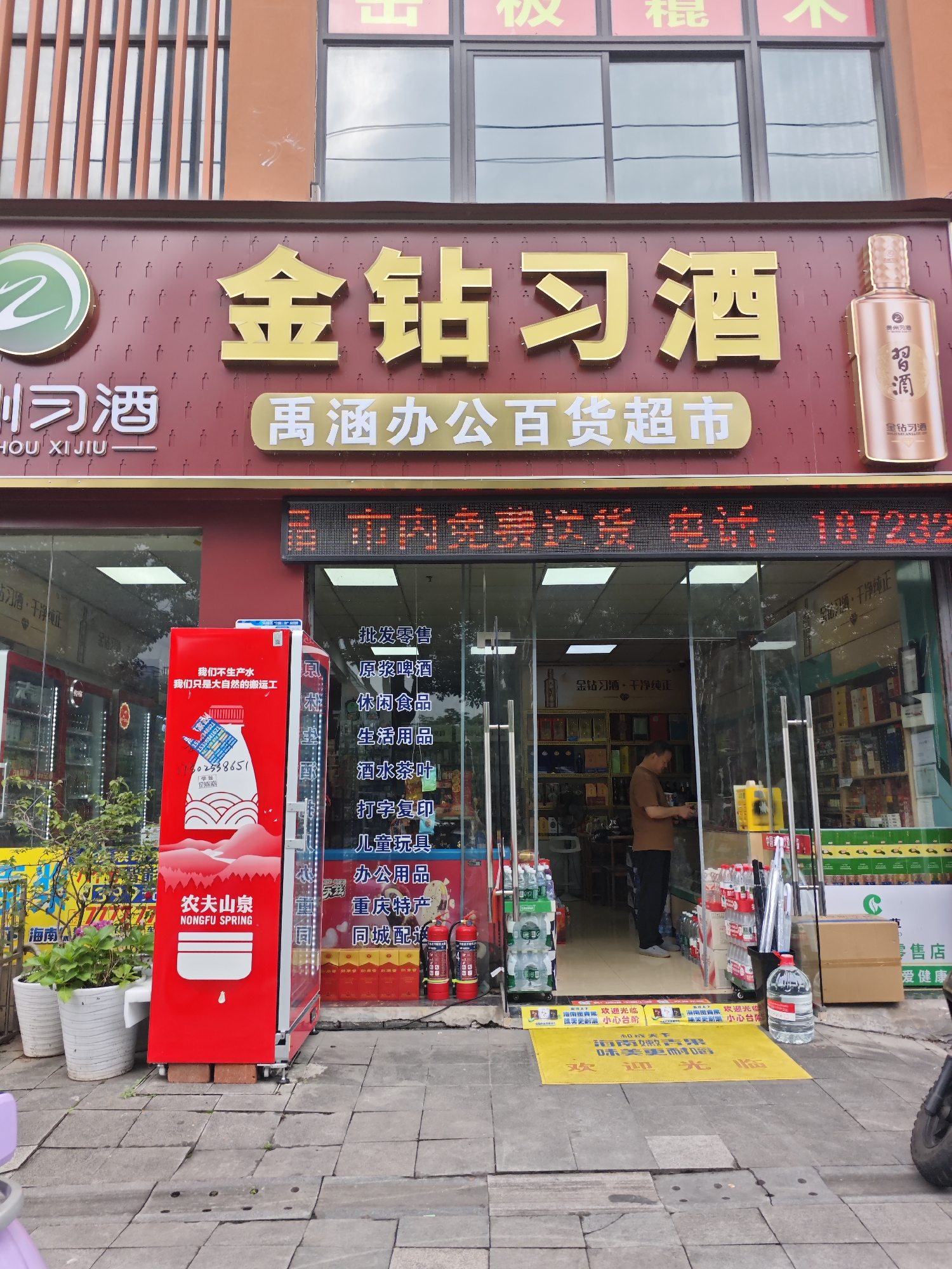 The image shows the storefront of a business in the city of Chongqing High-tech Zone, China. The business appears to be a convenience store or a liquor store, with signage indicating it sells alcohol and other goods. The facade features large, golden Chinese characters that translate to "Golden Diamond Liquor." Below this, smaller text reads "Gong Han General Merchandise Supermarket." A digital LED screen displays scrolling text that appears to be advertising or business information.
In front of the store, there is a red vending machine displaying advertisements for Nongfu Spring bottled water. Several potted plants are visible on the left side of the entrance. The store entrance is open, with a man in a brown shirt and dark pants visible inside, appearing to be interacting with merchandise. Shelves stocked with various products, including bottles of alcohol and other goods, are visible through the glass storefront. A yellow welcome mat with Chinese text is placed at the entrance. Fire extinguishers and other safety equipment are also present near the entrance. The street in front of the store is paved with concrete tiles.