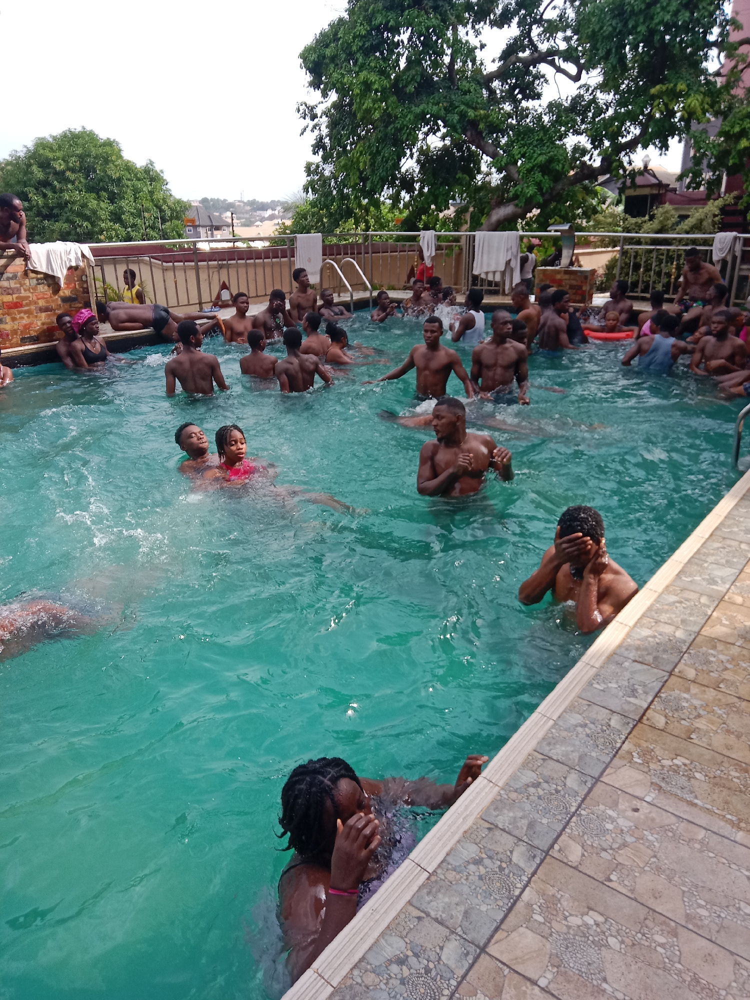 The image depicts a crowded outdoor swimming pool in Obosi, Nigeria. Many dark-skinned individuals, mostly young adults and some children, are in the water, some swimming and others standing or interacting. The pool is bordered by a tiled surface and a low brick wall with a metal railing. Several people are on the wall next to the pool. Towels are draped over the wall. Lush trees and vegetation surround the pool area, with buildings visible in the background. The water appears to be a turquoise color.