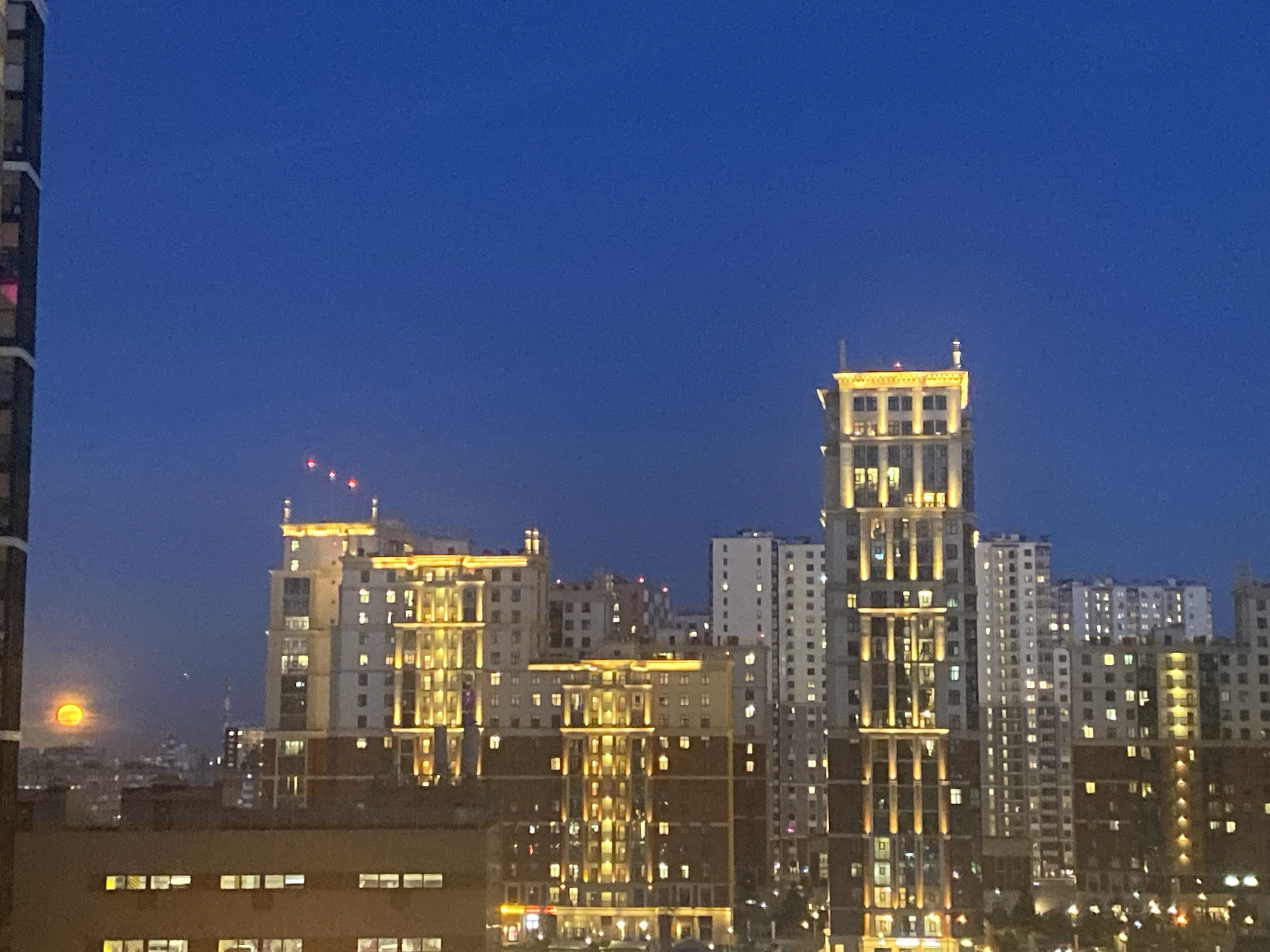 The image presents a panoramic view of a city at dusk or night, characterized by a clear, deep blue sky dominating the upper half. Numerous multi-story buildings of varying heights and architectural styles populate the cityscape. Many of these structures are illuminated, creating a vibrant urban glow.
In the foreground and mid-ground, several prominent buildings feature distinctive decorative lighting, particularly on their upper sections and along vertical architectural elements, casting a warm yellow light. These structures appear to have a more ornate, perhaps neo-classical, design. Interspersed among them are plainer, modern apartment blocks, identifiable by rows of glowing windows.
On the far left, a large, orange-hued full or nearly full moon is visible, appearing low in the sky and partially obscured by a nearby building or atmospheric haze. Three small, distinct red lights are visible in the dark blue sky above the buildings on the left side of the frame, possibly indicating an aircraft or distant communication towers. The very left edge of the image shows a partial view of another building, suggesting the photo was taken from an elevated position, perhaps from a window or balcony.
The scene depicts a static, quiet urban environment after sunset, with no human activity explicitly visible. The clear sky suggests good weather conditions. Given the provided context, the image captures a night view of Saint Petersburg, Russia. There is no visible text in the image.