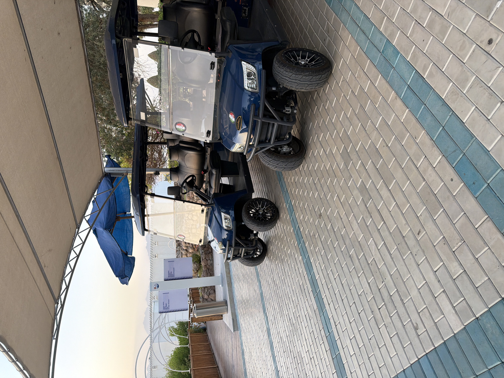 The image shows two dark blue golf carts parked under a tan awning in Sharm El Sheikh, Egypt. The golf carts are facing forward, with the one on the right in the foreground. Both carts have clear windshields and black seats. The golf cart in the foreground has a black bumper and custom black rims on its wheels. A small Italian flag emblem is visible on the windshield of the front golf cart. The ground is paved with light gray and blue rectangular tiles. In the background, there is a wooden walkway, some greenery, and a white structure with arches. The sky is a light blue, suggesting it is either early morning or late afternoon. The scene appears to be an outdoor area, possibly a resort or a golf course.
