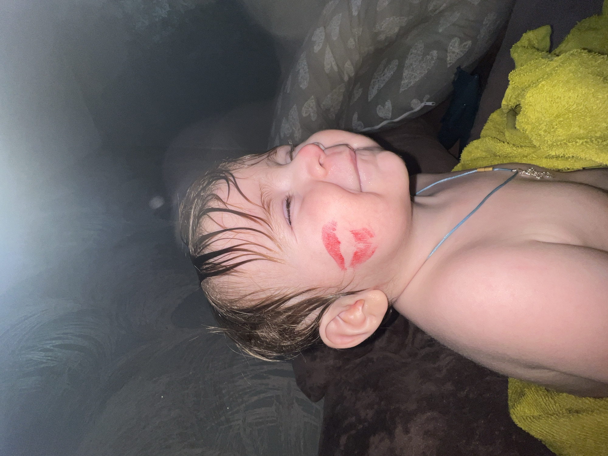 The scene depicts an infant or toddler in an indoor domestic environment. The specific location within Noviy, Russia, cannot be determined from the visual information. The child, with fair skin and short, wet-appearing light brown hair, is shown in a close-up, turned slightly to their right, with eyes closed and mouth forming a smile or grin, indicating amusement or contentment. A distinct red lipstick mark in the shape of lips is present on the child's left cheek. A thin light blue cord necklace with a small, indistinct metallic charm hangs around the child's neck. A bright yellow, textured towel is draped over the child's lower body. The background on the left consists of a dark brown, textured surface, likely upholstery. On the right, a light grey pillow with a repeating white heart and line pattern is visible, along with a white zipper. Further back, a dark blue or black fabric is partially observed. The lighting is subdued, with illumination highlighting the child's hair and skin.