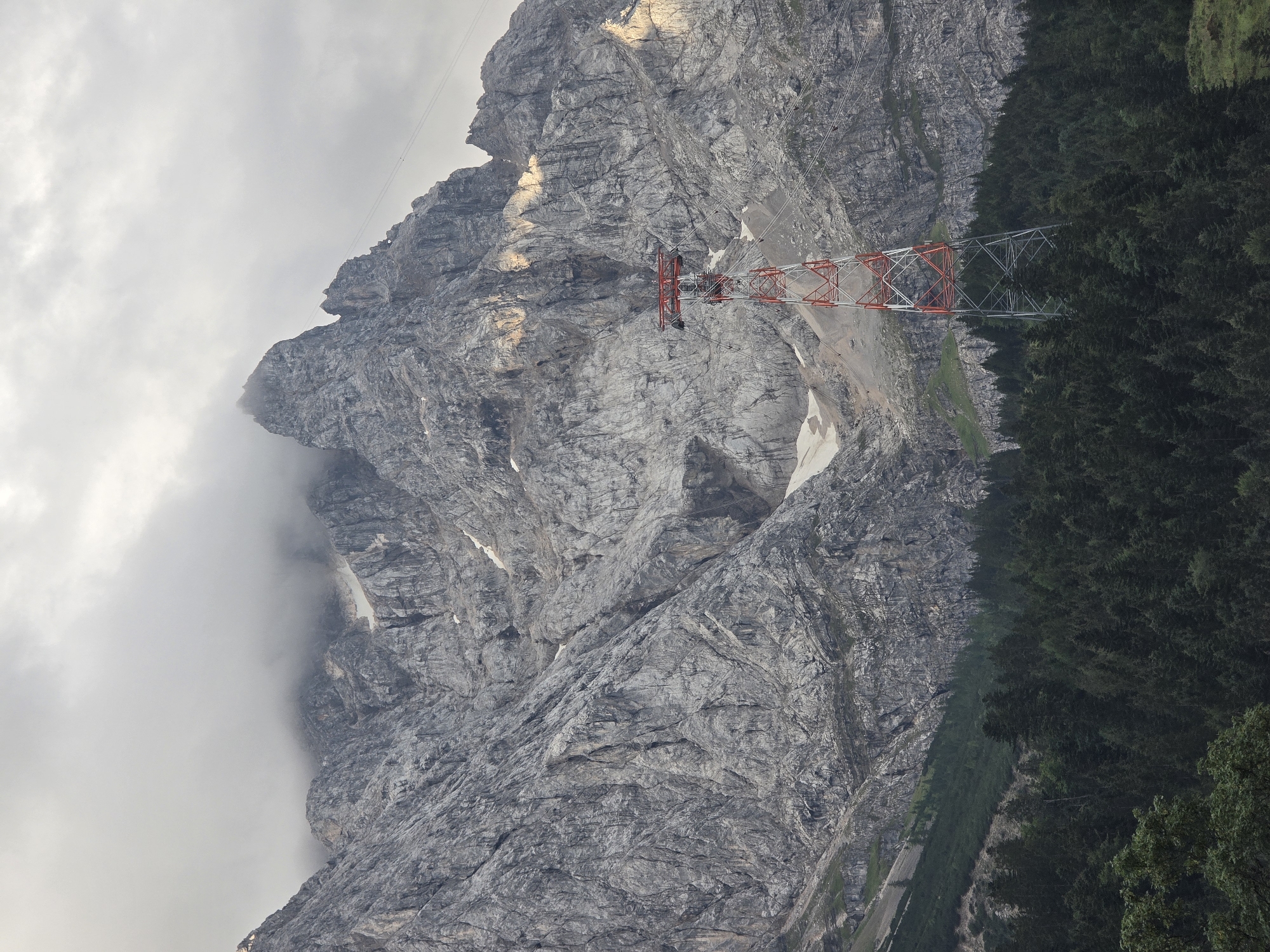The image captures a view of the Zugspitze mountain range in Grainau, Germany, with a red and white lattice tower for a cable car in the foreground. The mountain face is rugged and rocky, with patches of snow visible at higher elevations. Dense green coniferous forest covers the lower slopes. The sky is overcast with clouds, and a few rays of sunlight break through, illuminating parts of the mountain.