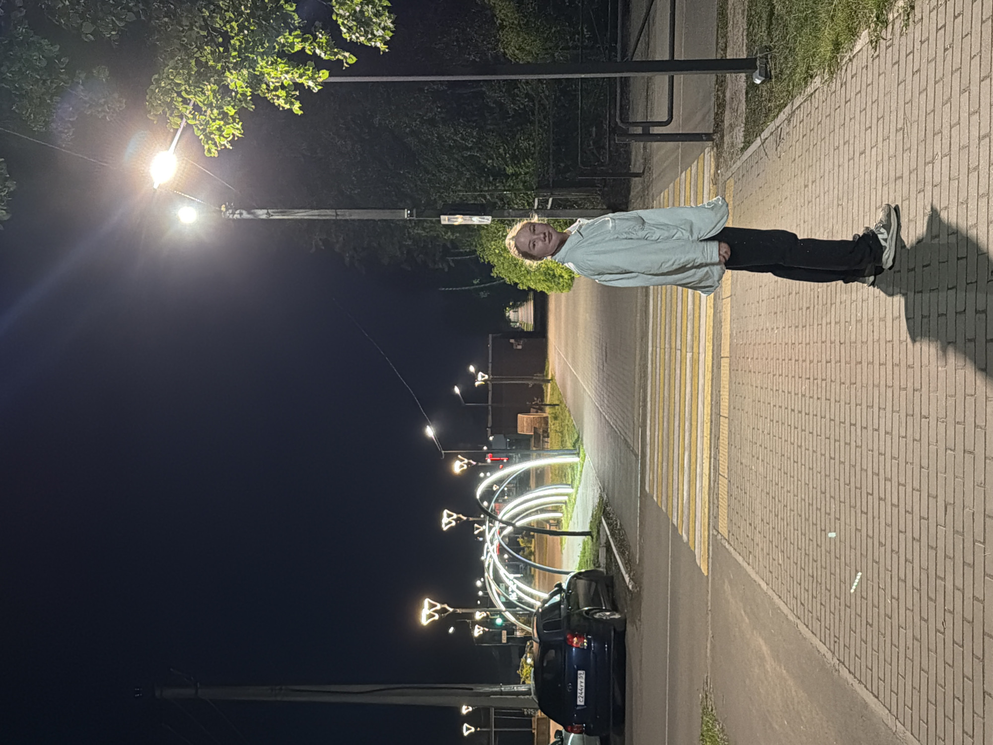 The image depicts an outdoor night scene featuring a young person standing on a sidewalk. The individual, likely a child or teenager, has light-colored hair and is wearing a light-colored jacket or shirt, dark pants, and light-colored sneakers. They are smiling and looking directly at the camera.
The setting is a street or pathway at night, illuminated by various artificial lights. To the right of the person (from the viewer's perspective) is a paved sidewalk with a brick pattern. To their left is a road with a clearly marked white and yellow striped crosswalk. A dark-colored sedan car is parked on the side of the road near the crosswalk; its license plate reads "C244YY59".
Multiple streetlights are visible throughout the scene, casting light and creating reflections. A very bright streetlight is positioned above and slightly behind the person. Further down the street, an artistic lighting installation consisting of several glowing white arches forms a tunnel-like structure. Smaller, decorative streetlights with distinct butterfly or bow-tie shaped fixtures are also part of this display. The background features dark trees and shrubbery against a dark night sky. The activity appears to be a casual moment captured at night, with the person posing for the camera.