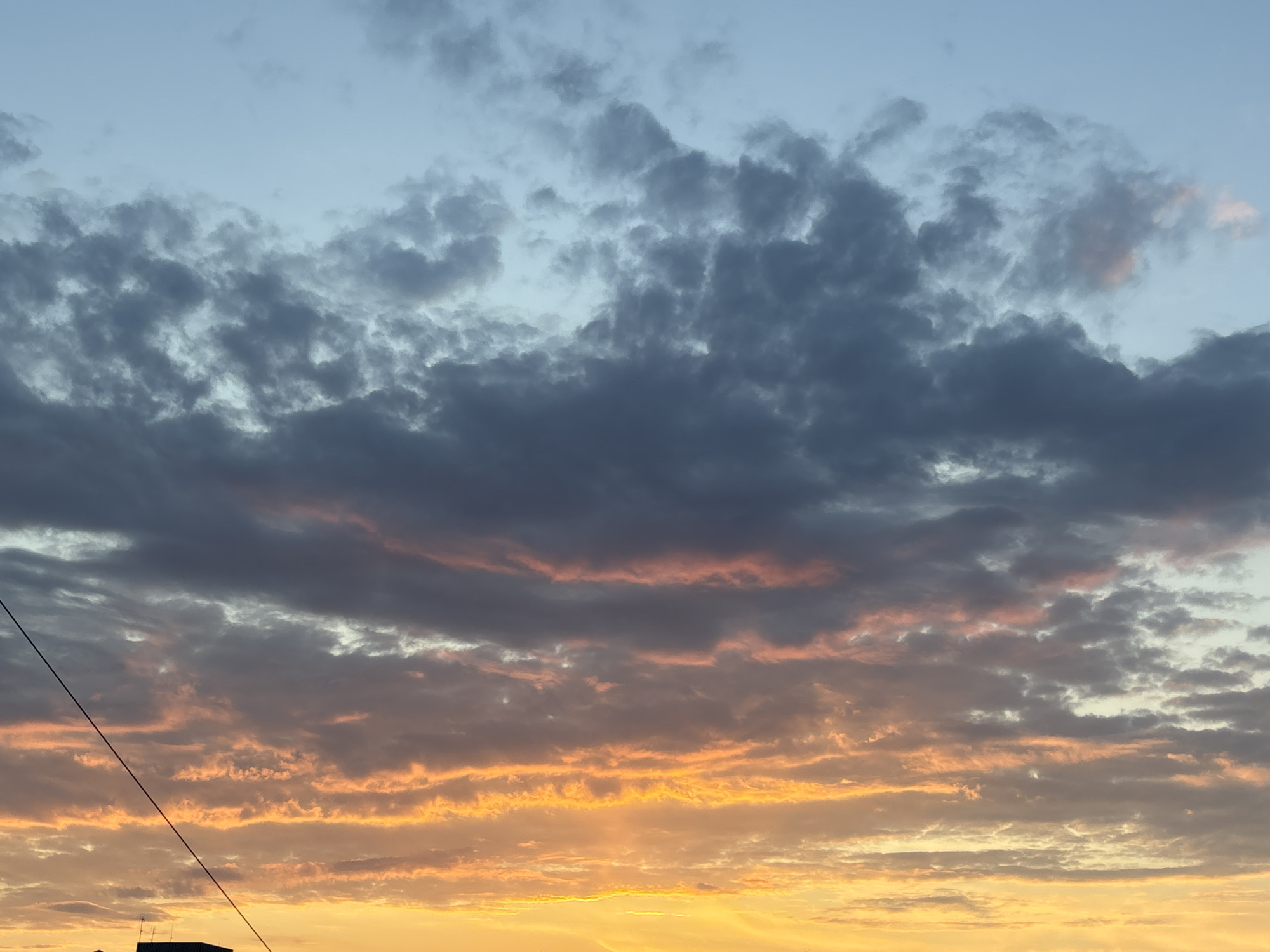 The image shows a sky filled with clouds at sunset. The upper portion of the sky is a pale blue, transitioning into dark gray clouds. Lower in the frame, the clouds are streaked with orange and yellow light, suggesting the sun is below the horizon. The lower portion of the image is a bright yellow with some clouds present. In the lower left corner, there is a glimpse of a dark building, and a thin black cable runs diagonally across the image. There is no indication this image was taken in Chaikovsky, Russia.