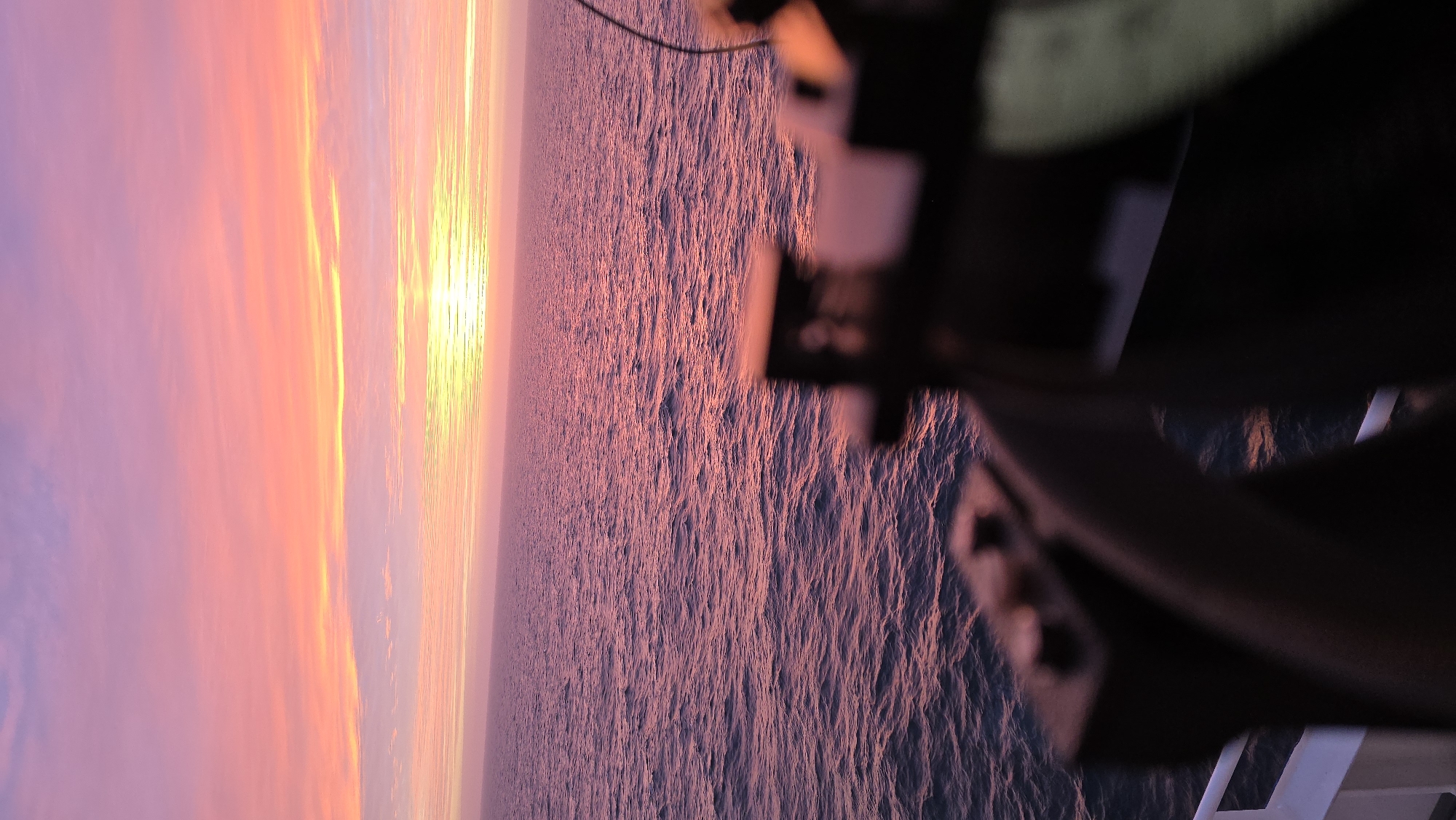 The image shows a sunset over water, possibly the ocean, as viewed from a ship or structure. The sky displays a gradient of colors, transitioning from orange and yellow near the horizon to pink and purple higher up. The water reflects these colors, creating a rippled texture. The foreground contains a blurry, dark structure, likely part of the ship or building, which obscures a portion of the scene. No people or specific location details are discernible from the image.