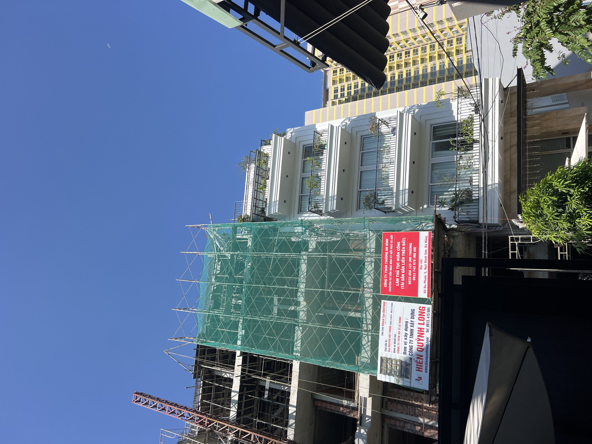 The image shows a construction site in a city with several buildings. In the foreground, a partially constructed building is covered in green safety netting. A large crane is visible extending from the top of the structure. On the side of the building, there are two signs with Vietnamese text and some images.
To the right of the construction site, a modern white building with multiple balconies is visible. These balconies are adorned with plants and railings. Further in the background, a tall yellow and grey building stands out against the clear blue sky. A faint crescent moon is visible in the sky in the upper right corner. The overall scene depicts an urban environment with ongoing development.