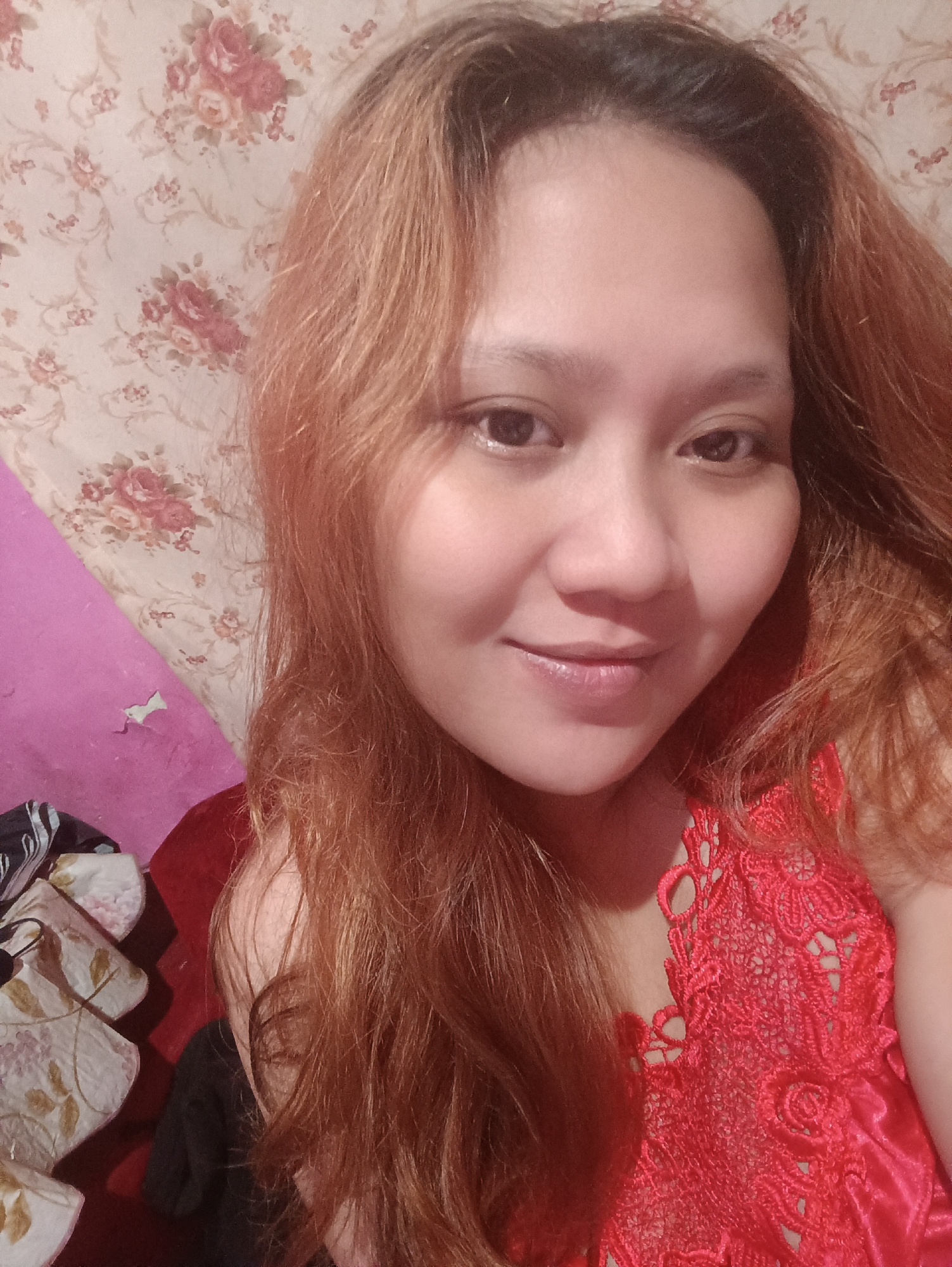 The image is a close-up, selfie-style photograph of a young woman with reddish-brown hair. She is looking directly at the camera with a slight smile. She is wearing a red lace garment. The background consists of floral wallpaper and a pink wall. The scene appears to be indoors, likely in a residential setting in Depok, Indonesia.