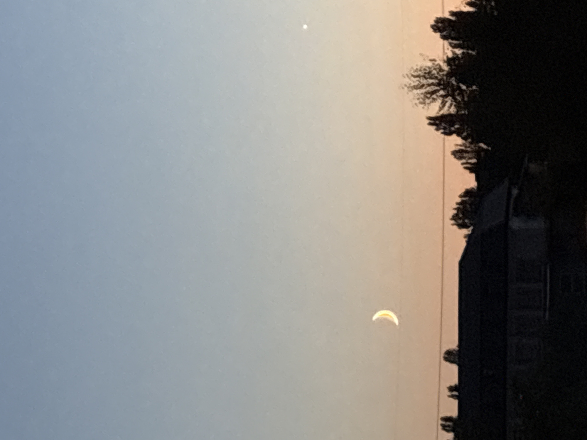 The image captures a serene twilight scene with a crescent moon hanging in the sky. A faint point of light, likely Venus, is also visible. Below the sky, a dark silhouette of trees and a building suggests an urban or suburban landscape. The building appears to be a house with a dark roof. The presence of trees suggests it might be a residential area with some greenery. The overall impression is one of a quiet evening, possibly in a residential district of Kyiv, Ukraine, where the transition from day to night is marked by celestial bodies and the silhouette of human habitation.