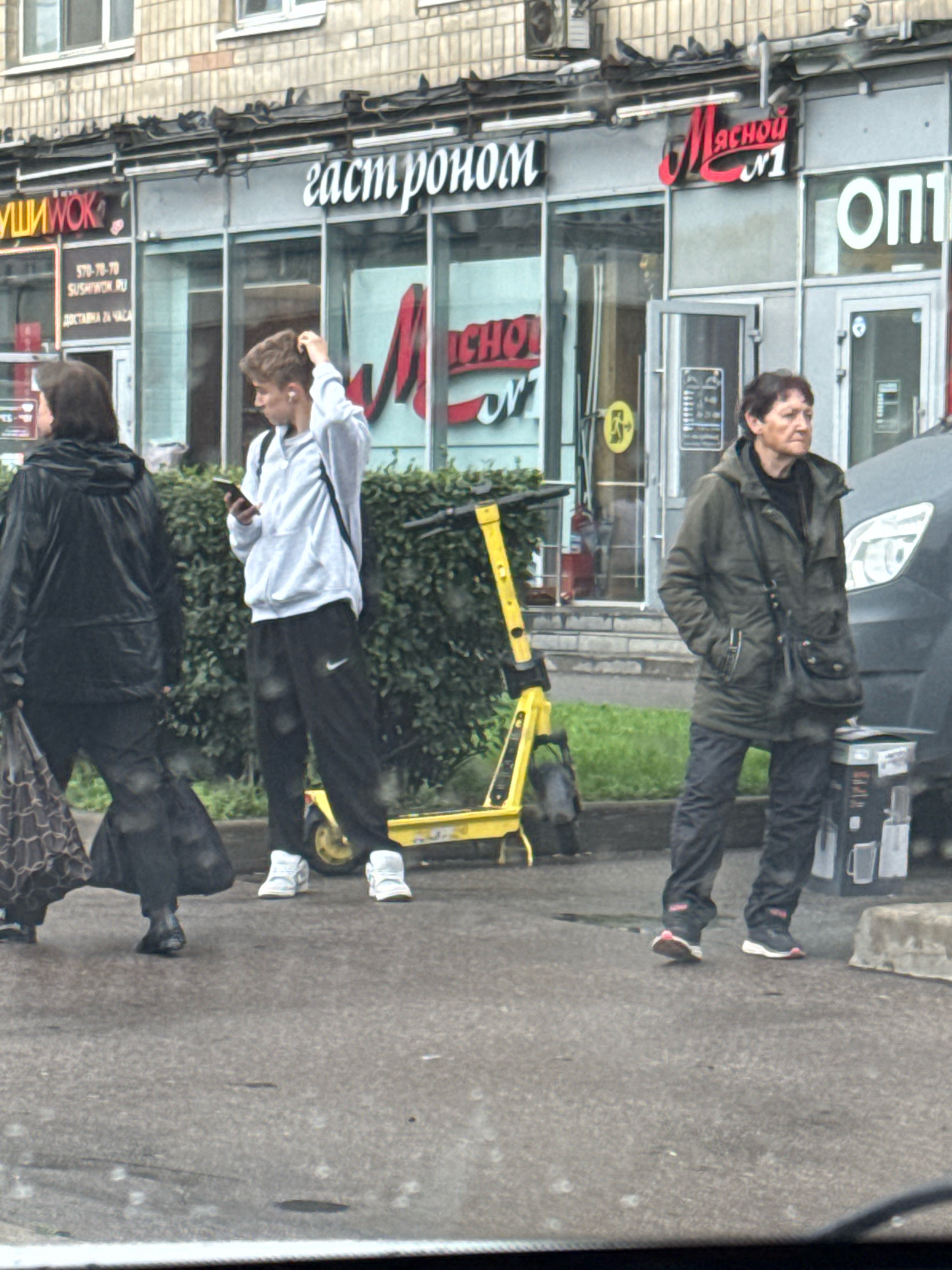 The image depicts a street scene in Saint Petersburg, Russia. In the foreground, a person in a black raincoat with a patterned bag and a black bag walks away from the camera. To their right, a young person in a light blue hoodie and black sweatpants with white sneakers is looking at their phone and scratching their head. Behind them, a bright yellow electric scooter stands upright. Further to the right, an adult wearing a dark green hooded jacket and dark pants with black and white sneakers stands with their hands in their pockets. They are looking off to the right side of the frame. In the background, the facade of a building features several storefronts with signage. One sign reads "ГАСТРОНОМ" (Gastronome), another has "МЯСНОЙ №1" (Meat No. 1), and another has "ОПТИ" (Opti). The pavement is wet, suggesting recent rain. A gray vehicle is partially visible on the far right.