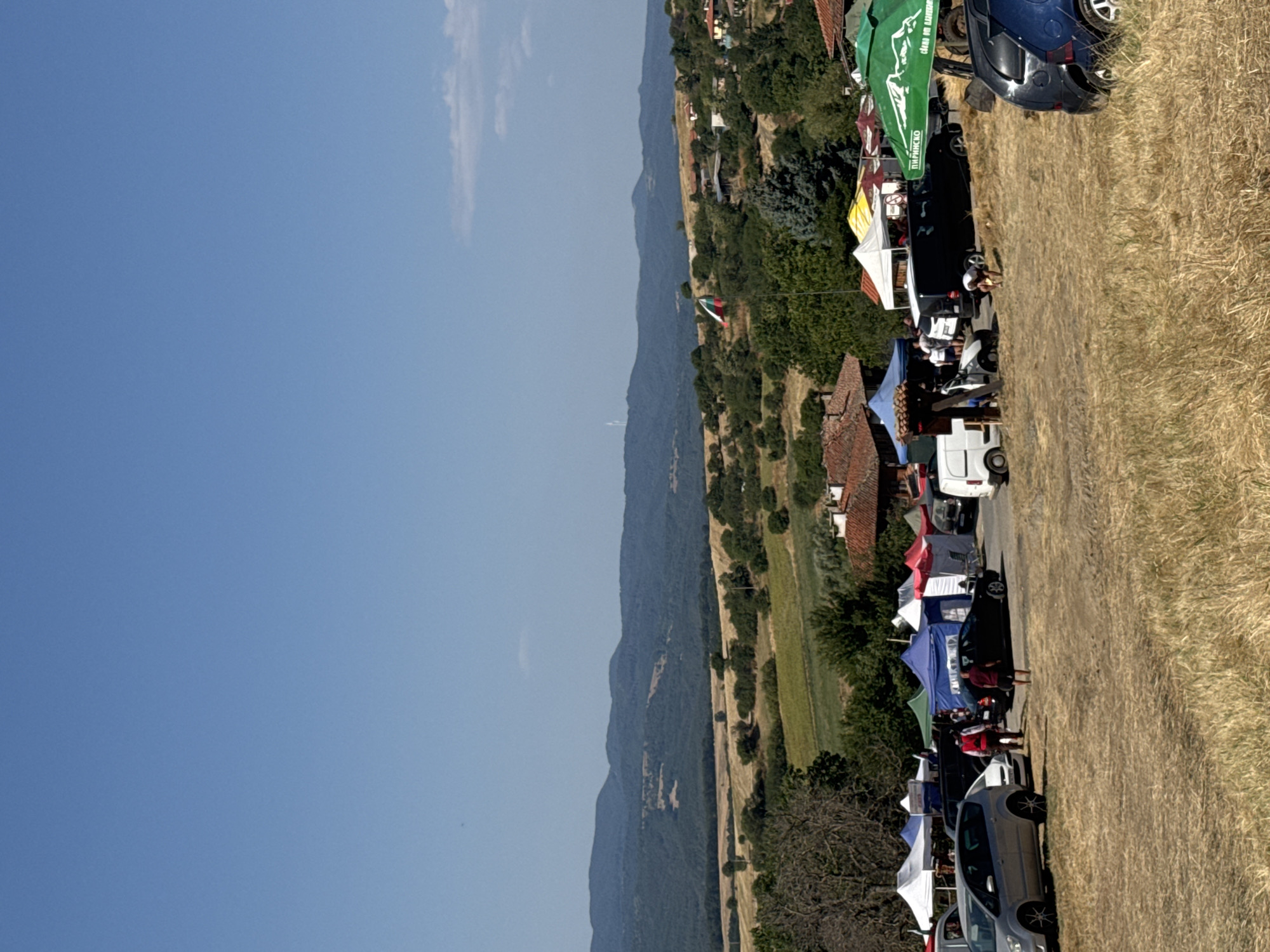 The image depicts an outdoor event, possibly a market or festival, set in a rural area with rolling hills and mountains in the background. The foreground is dominated by dry, golden grass, suggesting a summer or early autumn setting. Several tents and marquees are set up, some displaying colorful fabrics and signage. A Bulgarian flag is visible flying from a pole, hinting at the location being in Bulgaria.
In the middle ground, various vehicles are parked, including cars, vans, and what appears to be a small truck. People are seen interacting near the tents and vehicles, with some individuals walking and others engaged in activities. A person is bent down near a tent, possibly setting up or arranging items. The architecture of some buildings visible in the background, with red-tiled roofs, suggests a traditional village or town. The sky is clear and blue. The specific location within Zheravna, Bulgaria, is implied by the presence of the Bulgarian flag and the style of architecture.