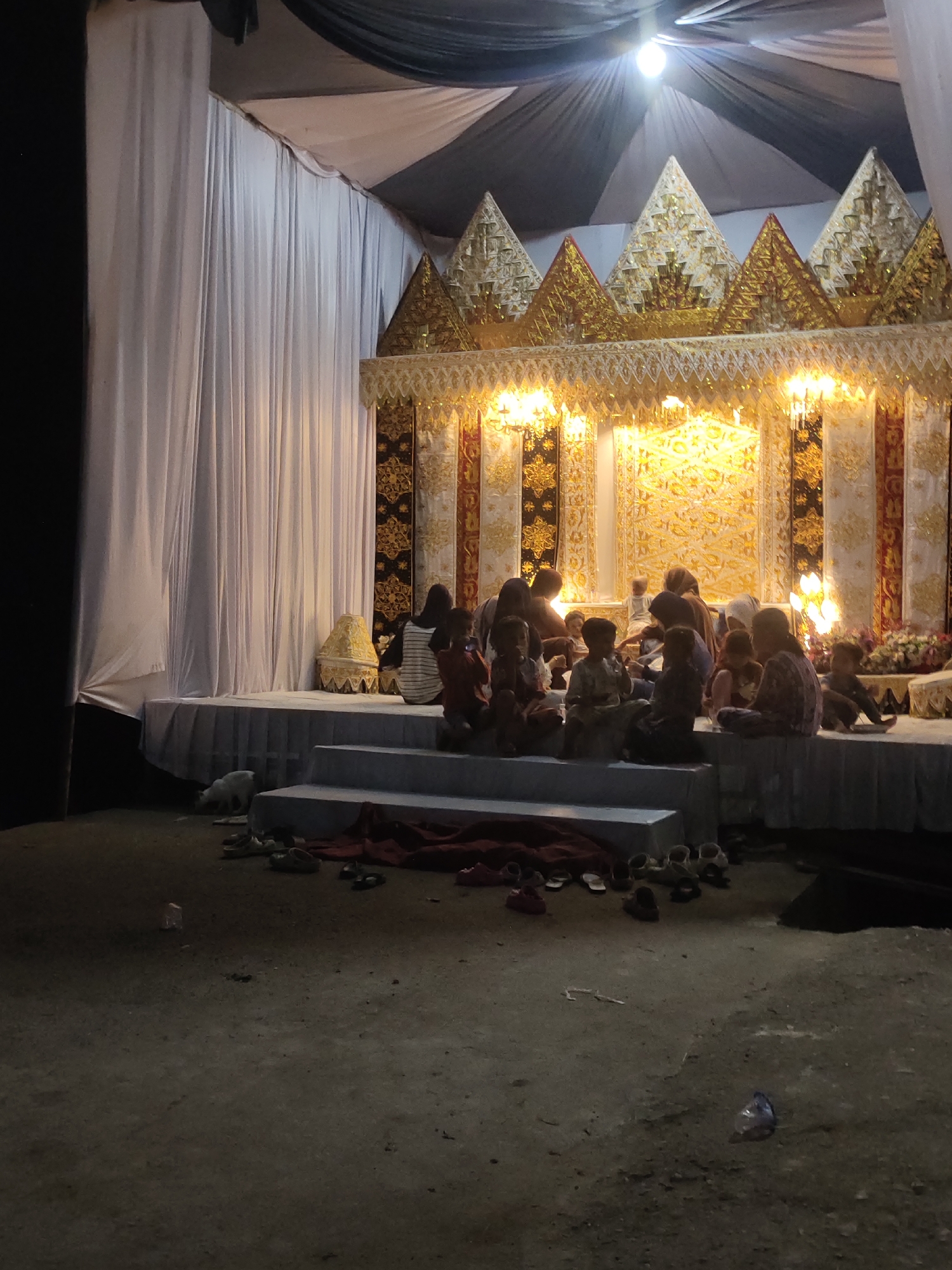 This image captures an evening or night gathering around an elaborately decorated stage, likely outdoors, in Lhokseumawe, Indonesia. The scene is illuminated by artificial lights emanating from and above the stage, casting the foreground in dim light.
The focal point is a multi-tiered stage adorned with intricate, golden traditional Indonesian decorations featuring pointed peaks and patterned fabrics, reminiscent of a *pelaminan* (wedding stage). White drapes form a backdrop and cover the ceiling, with some black drapes also visible. Bright spotlights shine down from above the stage. On the left side of the stage, a large white drape hangs vertically.
A group of people, primarily children and some adults, are seated on the edge of the stage platform and the steps leading up to it. They appear casually dressed and are facing towards the stage's center, seemingly observing an event or performance. Below the stage, a white cat is visible near the left edge. Numerous pairs of shoes are scattered on the ground near the steps, indicating that people removed them before going onto the platform. A piece of red fabric or a blanket is also seen on the steps, and a small plastic item lies on the dirt ground in the foreground.
The atmosphere suggests a community event, possibly a wedding or another ceremonial gathering, taking place in the late hours. No visible text is present in the image.