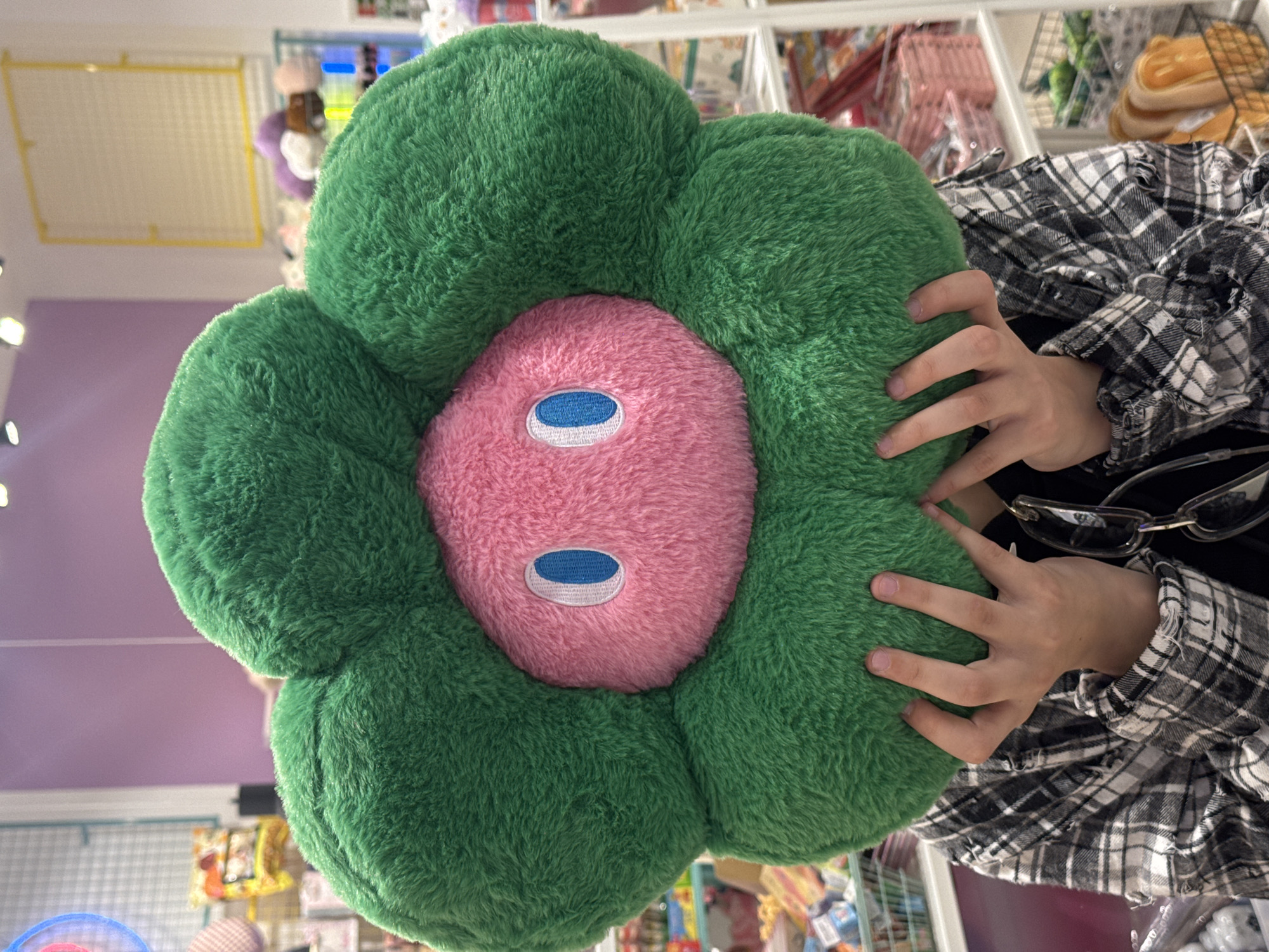 A close-up shows a plush flower pillow held by a person in a black and white plaid shirt. The pillow features green petals surrounding a pink center with blue and white circular accents. The background shows a store environment with shelves containing various items. The city of Moscow, Russia is not discernible from the provided image.