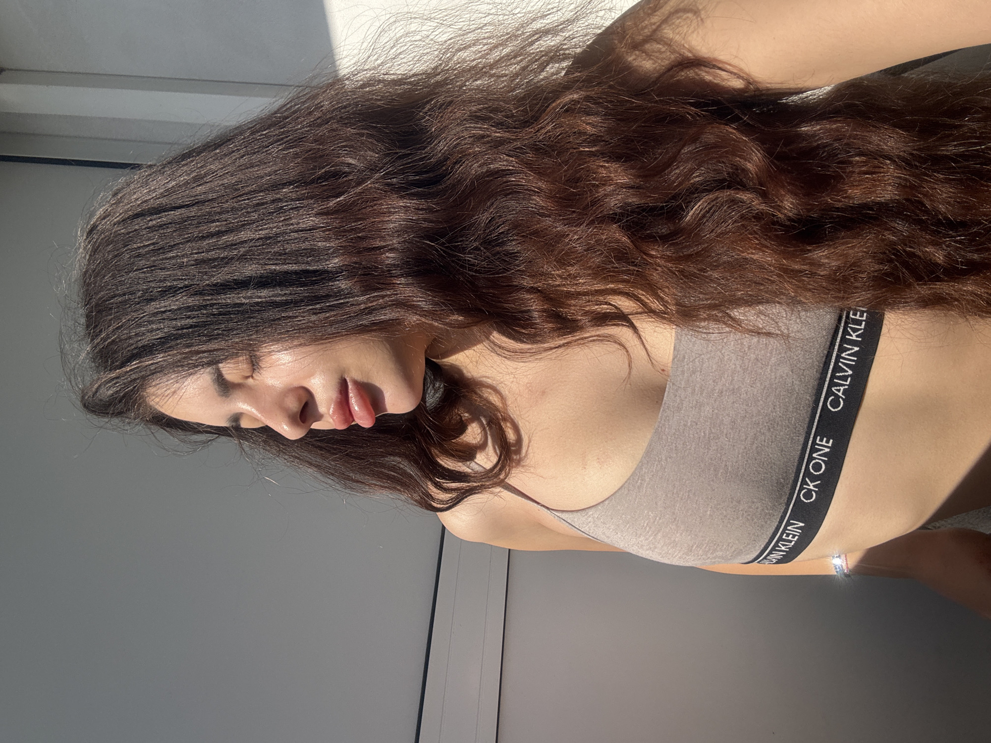 A young woman is photographed in what appears to be an indoor setting. She is wearing a Calvin Klein sports bra. Her long, dark hair is styled in loose waves. The environment seems to be bathed in natural light. There is nothing that indicates that the image was taken in the city of Plovdiv, Bulgaria.
