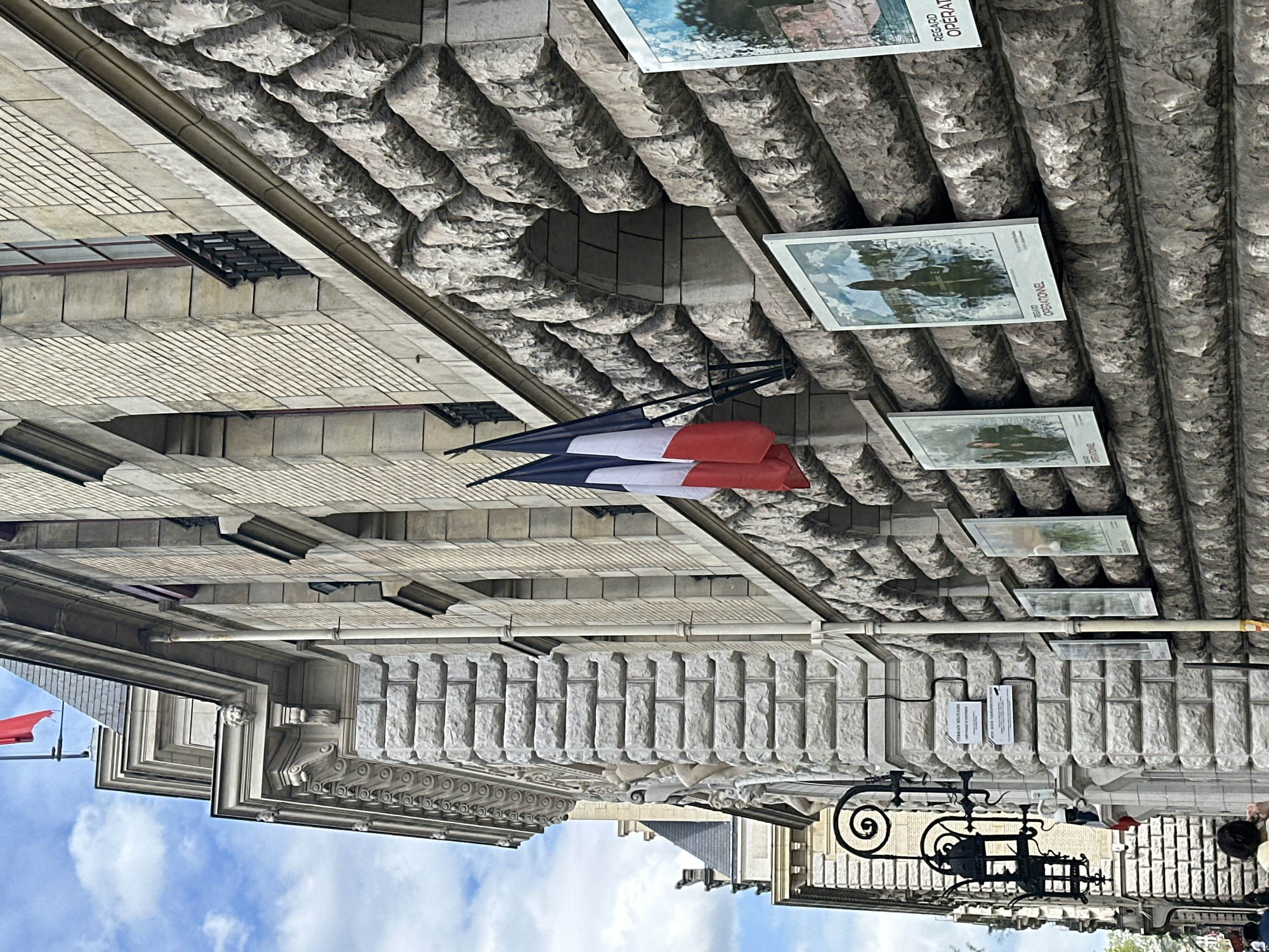 An outdoor scene in Paris, France, captures a grand stone building under a bright, partly cloudy sky. The building exhibits intricate classical architecture, featuring rough-hewn, textured stone blocks (rustication) on its lower levels, contrasted with smoother stonework and decorative cornices higher up. A French flag, displaying vertical blue, white, and red stripes, hangs prominently from a black bracket affixed to the building.
Below the flag, a series of framed posters or signs are attached to the stone wall, forming an outdoor exhibition. These posters primarily show images, some depicting landscapes or figures, and prominently feature the French text "REGARD OPÉRATIONNEL." One sign also includes "TERRAIN MILITAIRE" and "INFORMATIONS D'EXPOSITION." The arrangement of the posters suggests a curated display for public viewing.
In the lower right, a decorative wrought-iron railing or gate with scrollwork is visible. A person's head with dark hair is partially seen behind this structure. The lighting indicates it is daytime, likely sunny or partly cloudy, creating clear shadows and highlighting the details of the stone facade. The overall impression is one of a historic urban environment with cultural elements on display.