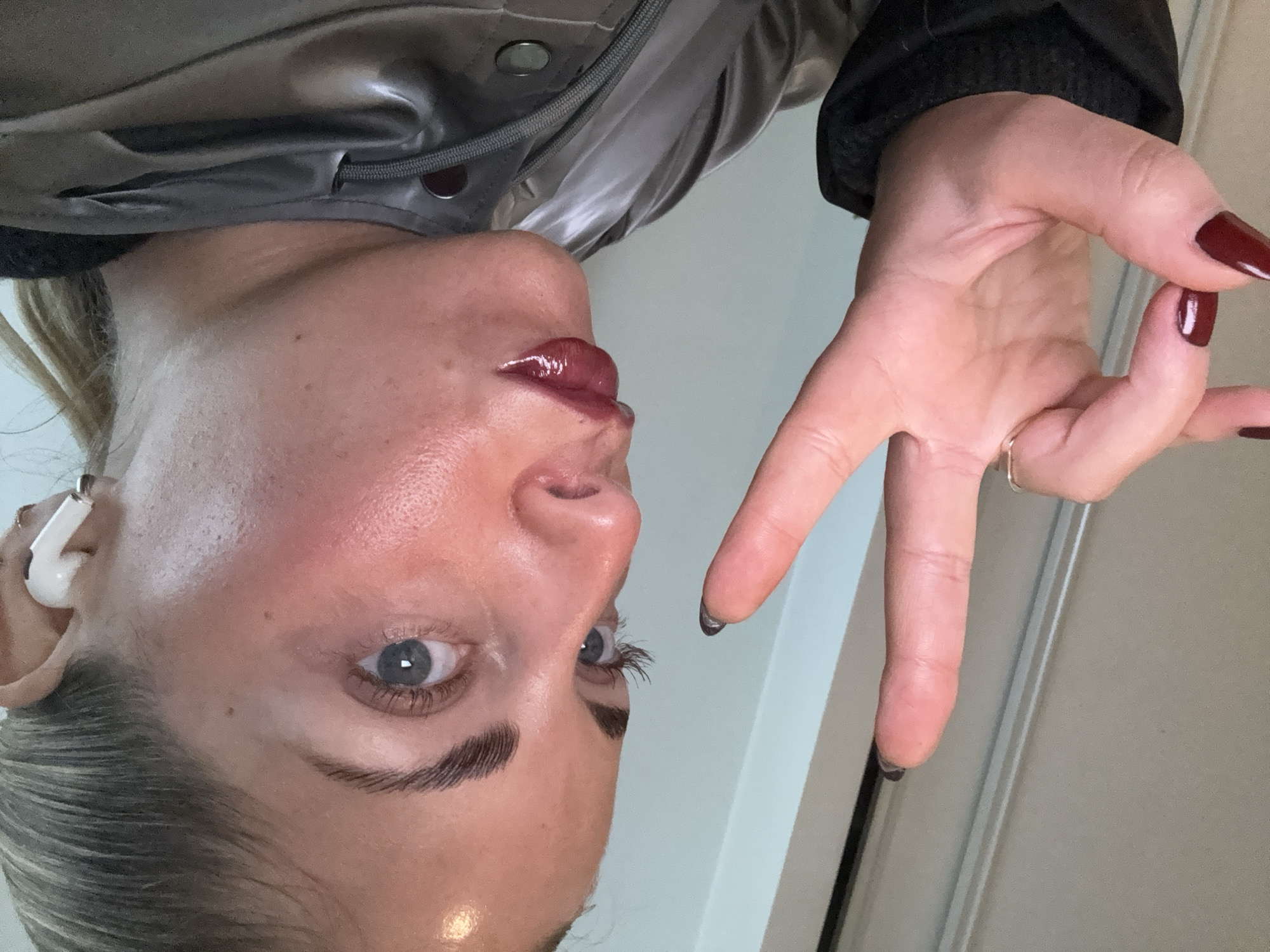 A woman's face is prominently featured in the image. She has fair skin, light-colored hair, and blue eyes. She is wearing an earpiece in her left ear and has glossy red lipstick on. Her eyebrows are neatly shaped. Her right hand is raised, displaying dark red nail polish. She is wearing a metallic jacket and a ring on her right hand. The background is a plain door. The location is not discernible from the image.