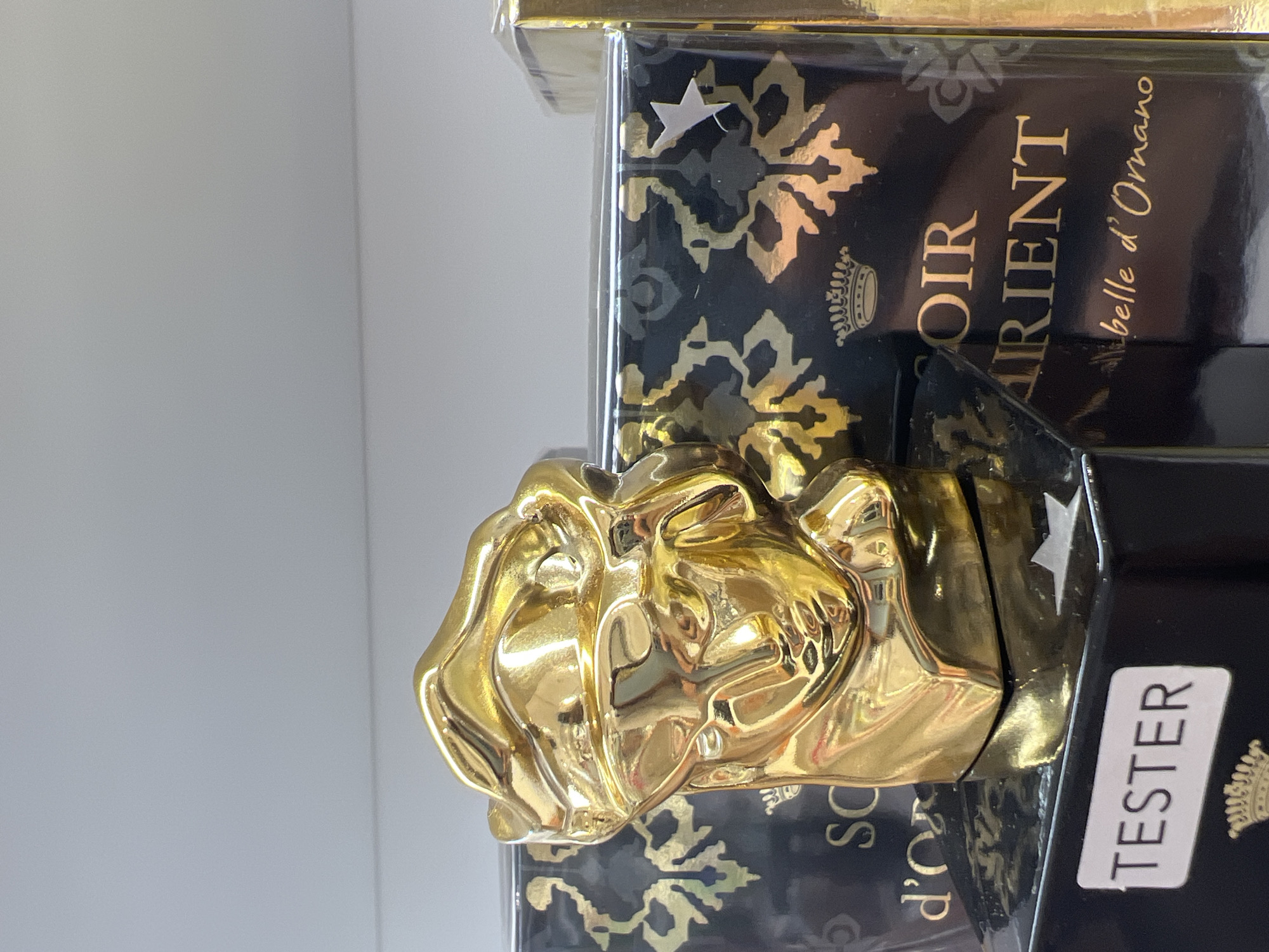 The image shows a golden bust of a person with abstract features, possibly representing a mask or a stylized head, placed on top of a black box. The box has ornate gold patterns and text. The text "TESTER" is visible on a white label affixed to the front of the black box. In the background, there are other boxes with similar gold patterns. The scene appears to be indoors, possibly in a retail or display setting, with a white shelf or wall in the upper portion of the image. The lighting is bright and reflective, highlighting the golden sheen of the bust and the metallic elements on the boxes. There are no people visible in the image. The specific location within Sofia, Bulgaria is not discernible from the image content.