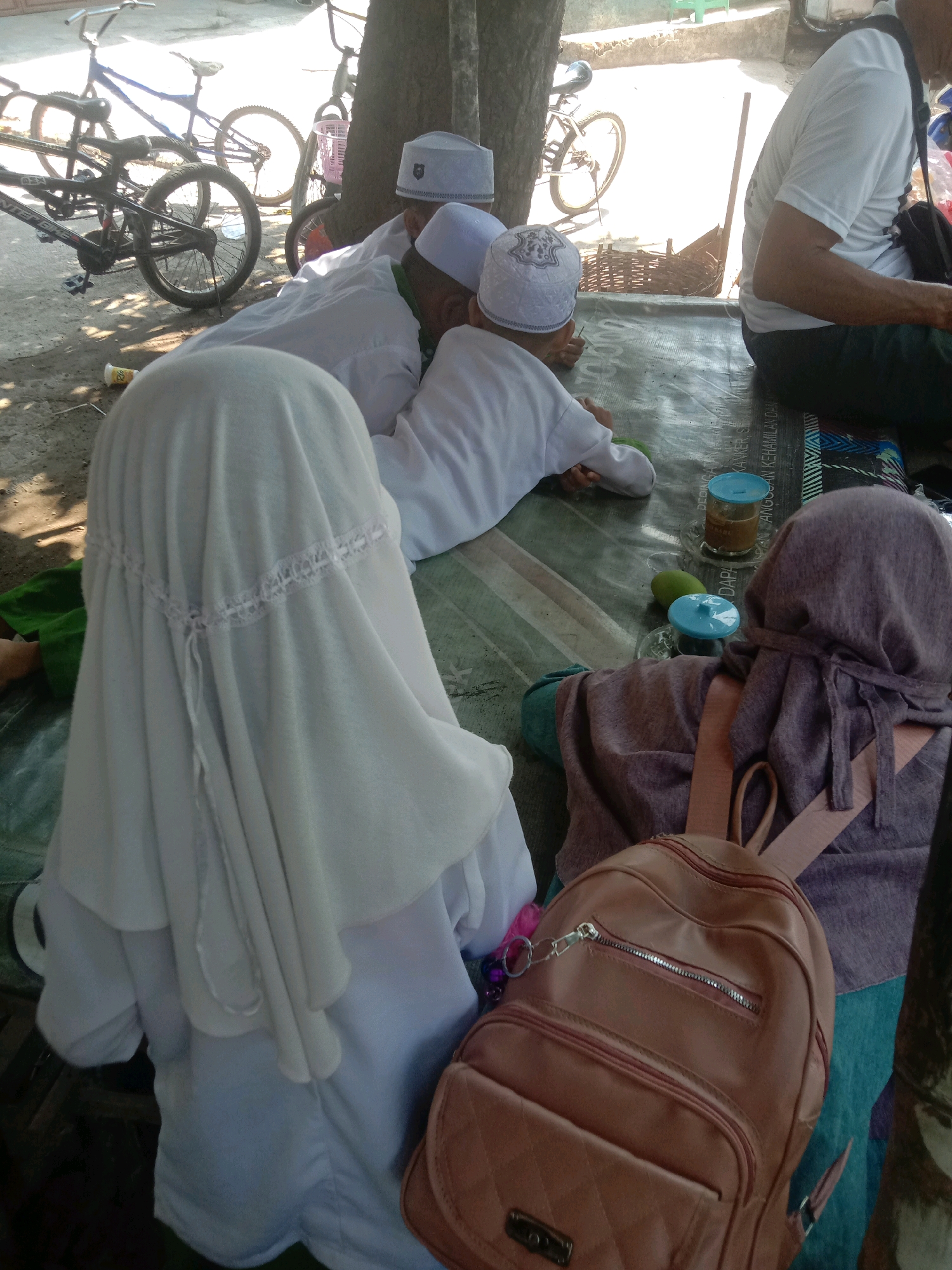 In the city of Ngempit, Indonesia, on a sunny day, three children are sitting on a mat, presumably for a religious lesson. Two children are wearing white Islamic skullcaps and white clothing, indicating they are likely boys engaged in religious studies. One of the boys appears to be a young child, possibly a toddler, observing something with interest. The third child, facing away from the camera, is wearing a white hijab and a white garment, suggesting they are a girl. Another child, also facing away, is wearing a purple hijab and a purple garment, with a pink backpack slung over their shoulders. The children are seated on a plastic mat outdoors, with bicycles visible in the background. A man in a white t-shirt and dark pants can be seen sitting to the right of the frame, facing the children, with a backpack on his lap. Several objects are scattered on the mat, including a glass jar with a blue lid, a green fruit (possibly a mango), and a blue cup with a lid. The scene suggests a relaxed outdoor learning or gathering environment.