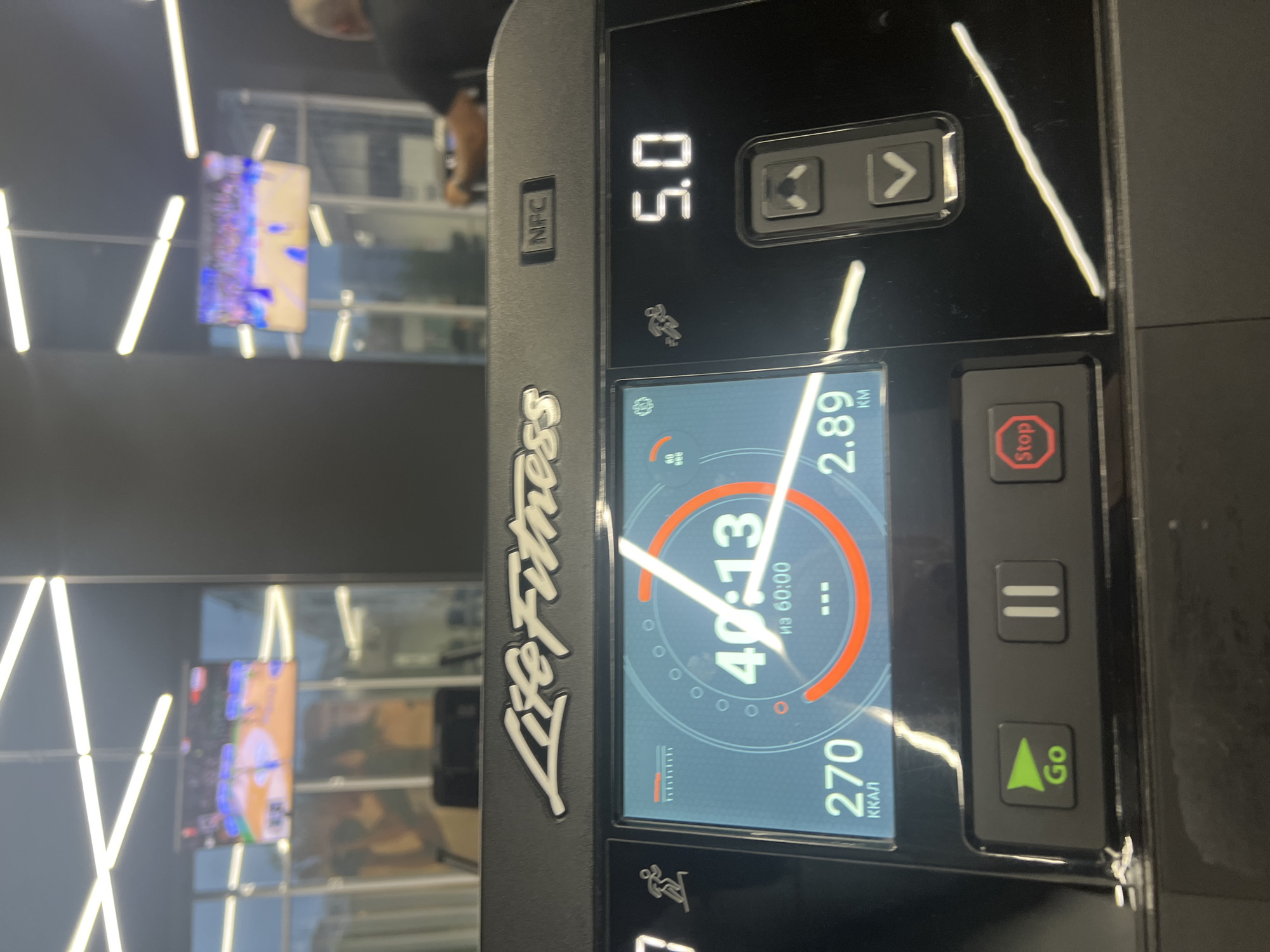 The image shows the control panel of a Life Fitness treadmill. The display indicates a workout time of 40 minutes and 13 seconds out of 60 minutes, a distance of 2.89 kilometers, and 270 kilocalories burned. The speed is set to 5.0. Buttons for "Go" and "Stop" are visible, as well as a pause button. The background includes a wall with linear lighting fixtures and blurry images of screens.