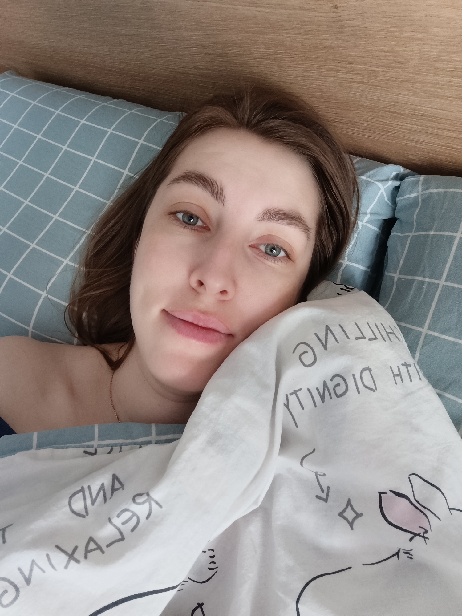 The image captures a female individual lying in bed in Chaikovsky, Russia. The subject has fair skin, light brown hair, and light blue-green eyes, looking directly at the viewer with a slight smile. Her left shoulder and upper chest are bare, displaying a thin gold chain around her neck. She rests against a light blue pillow featuring a white grid pattern. A white duvet covers the lower portion of her body and part of her right shoulder, adorned with mirrored grey and black text that, when unmirrored, reads "RELAX AND CHILLING WITH DIGNITY." The duvet also features a stylized black outline drawing of a figure that resembles a rabbit's head with a pink inner ear, along with abstract shapes like a diamond and arrows. The background consists of a light brown wooden headboard with horizontal grain. The scene is an indoor, well-lit bedroom environment.