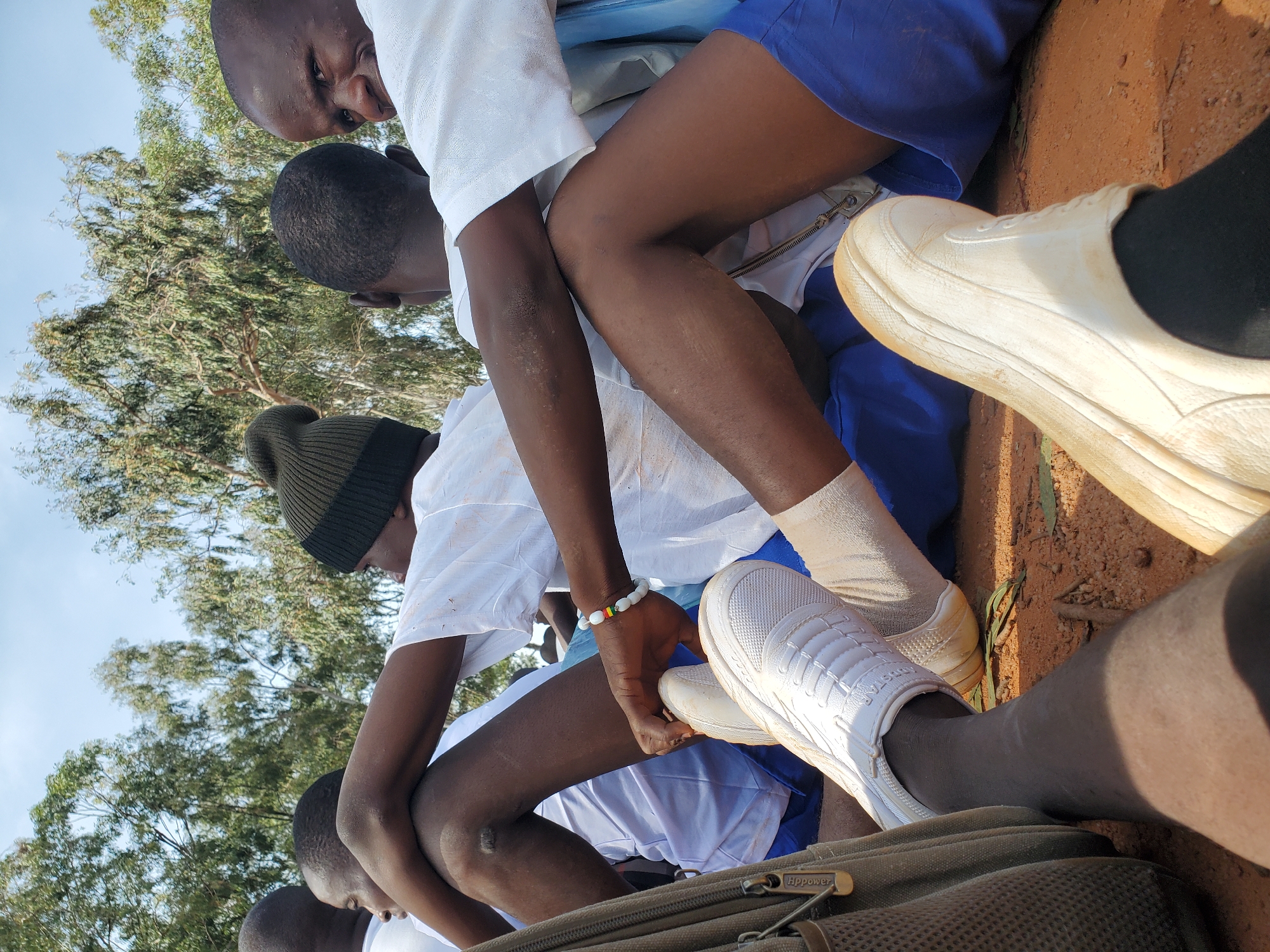 In Barakin Rafin Gora, Nigeria, a group of young people, likely students, are seated outdoors. They appear to be gathered in a relaxed, informal setting, possibly during a break or for an event. The individuals are seen from the waist down, with their legs crossed and arms resting on each other, suggesting a sense of camaraderie.
The individuals are wearing school uniforms, consisting of white shirts and blue shorts or skirts. Some have on white sneakers and socks. One person is wearing a dark knit hat. In the foreground, a backpack with a zipper is visible.
The ground is covered in reddish-brown dirt, with some dry leaves scattered around. In the background, several trees with green foliage are visible against a light blue sky. The overall impression is one of a bright, outdoor environment, typical of a school or community gathering space in Nigeria.