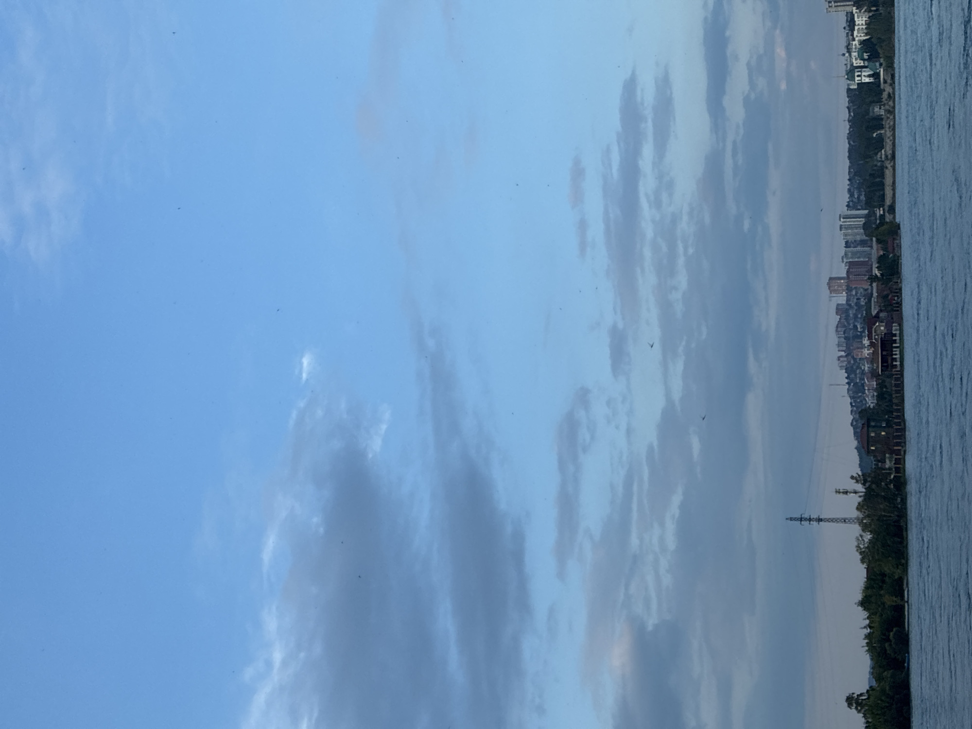 An aerial view shows a panoramic scene of a city skyline along a body of water. The sky is a clear blue with scattered clouds. In the foreground, the water is calm and reflects the sky. A distant city skyline features a mix of residential and commercial buildings, with some taller structures reaching towards the sky. Green trees and vegetation are interspersed with the buildings, creating a natural border between the city and the water. The overall impression is one of a peaceful urban landscape. The specific location in Irkutsk, Russia, is the Angara River embankment, showcasing the city's waterfront development and its connection to the natural environment.