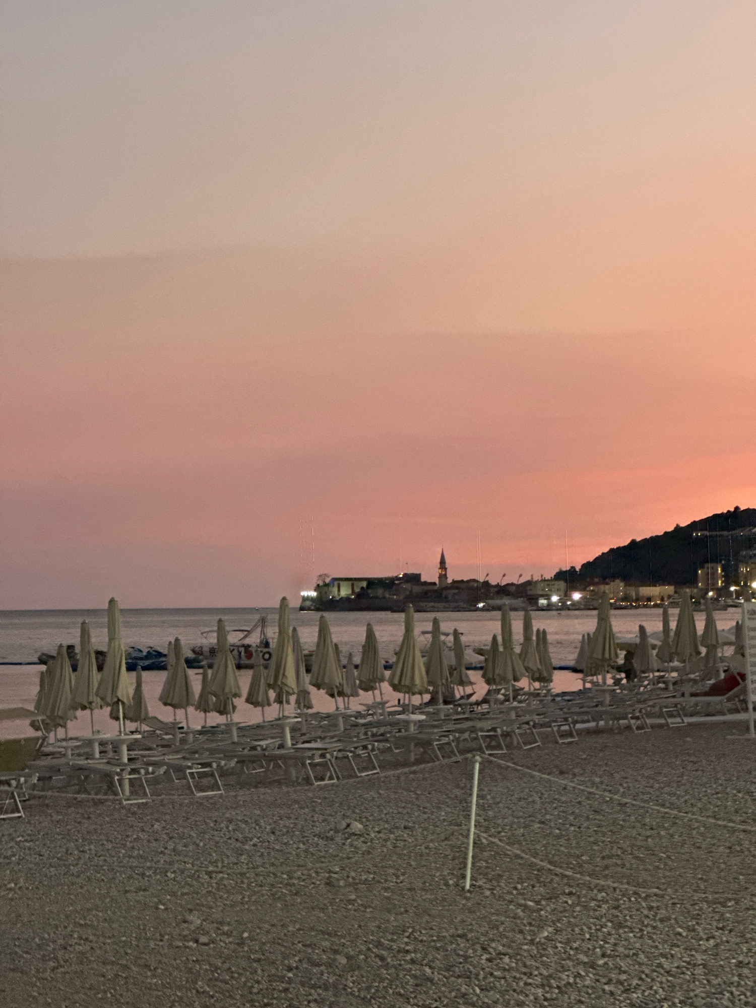The image captures a tranquil scene on a pebble beach in Budva, Montenegro, during twilight. The sky is a soft gradient of pink and orange, transitioning to a lighter hue towards the horizon, indicating either sunrise or sunset. In the foreground, rows of closed beach umbrellas and empty lounge chairs are neatly arranged, suggesting the end of a day or the anticipation of a new one. The beach itself is covered in small, grey pebbles.
Beyond the beach, the calm sea stretches towards a distant coastline. The cityscape of Budva is visible across the water, characterized by buildings with warm lights illuminating them against the darkening sky. A prominent church tower stands out amongst the structures, adding a touch of historical architecture to the scene. A densely vegetated hill rises behind the city, silhouetted against the twilight sky. The water is calm with a few boats and possibly jet skis visible, bobbing gently. The overall atmosphere is serene and peaceful, with no people actively present on the beach in the immediate foreground.