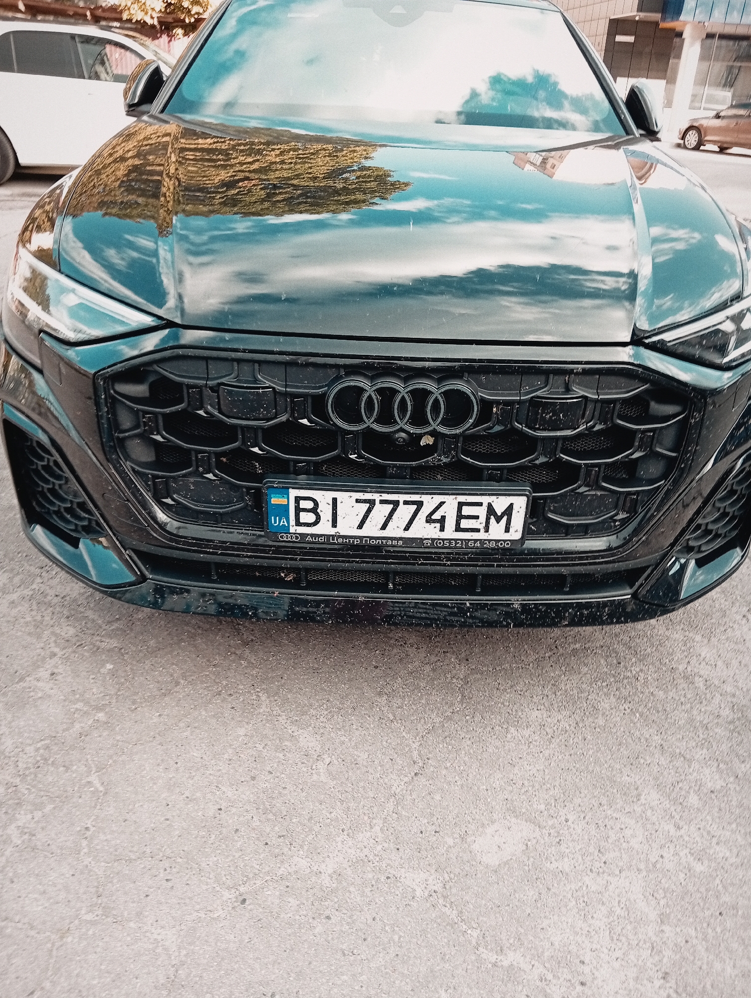 This media file shows the front of a dark-colored Audi Q8 parked on a paved surface. The car's grille, headlights, and license plate are clearly visible. The license plate displays "BI 7774 EM" and includes "UA" in a blue and yellow square, indicating Ukraine. Below the license plate, text reads "Audi Центр Полтава" and a phone number "(0532) 64 28 00". The car's hood reflects the sky and surrounding buildings, suggesting it is daytime with a partly cloudy sky. The pavement appears slightly wet or dirty. Another car, possibly white, is partially visible to the left, and a building facade is seen in the background. There are no people visible in the media file. The overall impression is that of a static shot of a car in an urban or suburban setting.