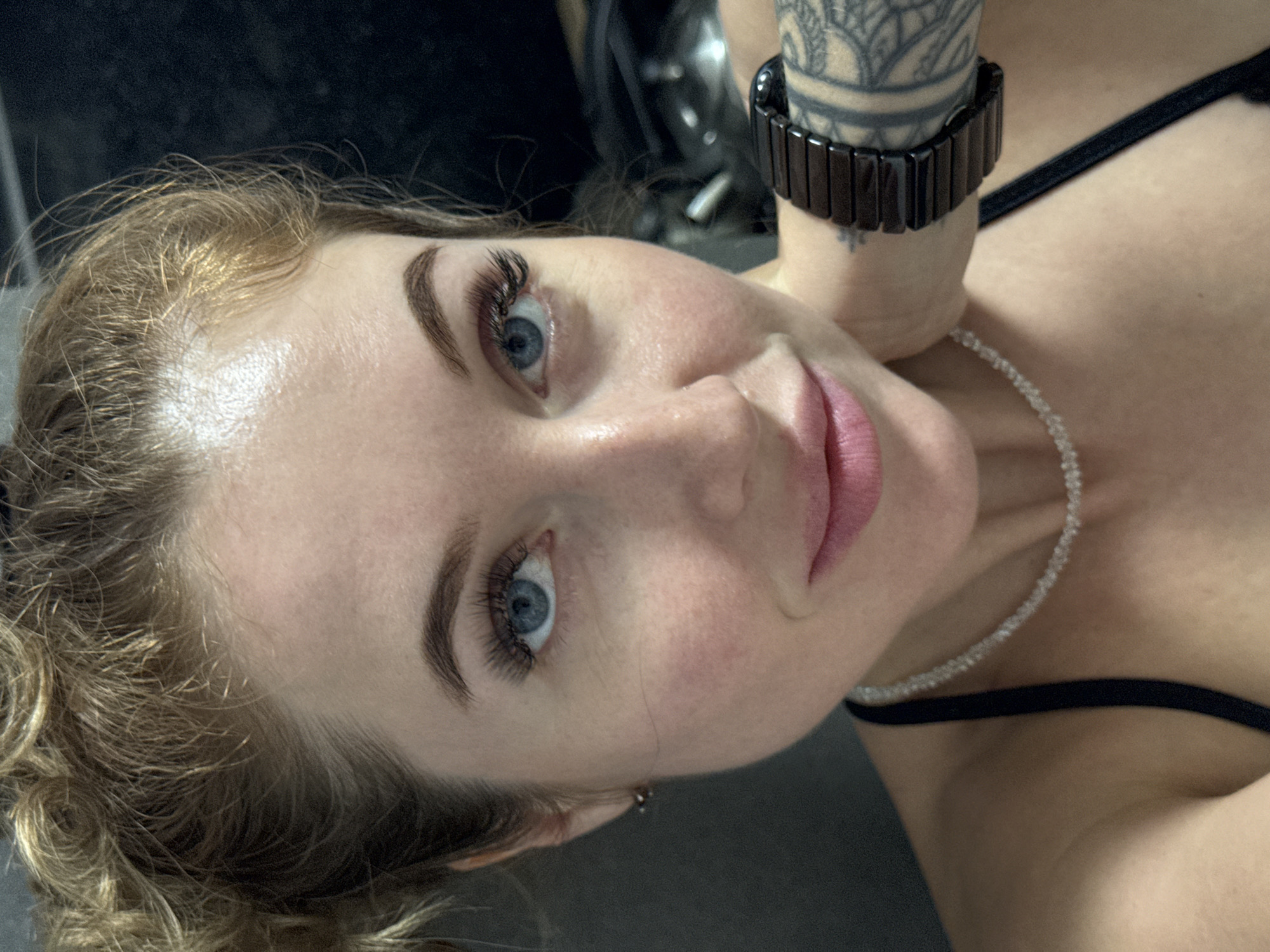 A close-up, angled portrait captures a fair-skinned woman with blue eyes. She has blonde, wavy hair and visible makeup, including defined eyebrows and eyelash extensions. She wears a black, possibly ceramic, wristwatch on her left wrist, which is adorned with a tattoo. A sparkling necklace encircles her neck. A thin black strap of clothing is partially visible on her shoulder. The background is indistinct. There is no evidence to determine the image was taken in the city of Chaikovsky, Russia.