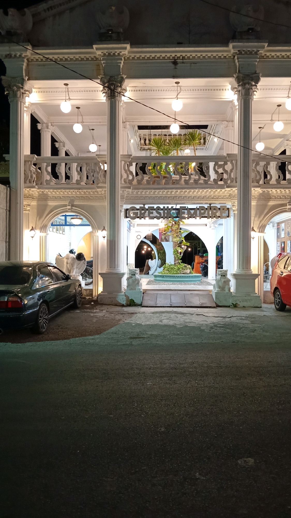 The image depicts the entrance of "Gresik Empire" in the city of Suci, Indonesia, at night. The building features a grand, white facade with classical architectural elements, including large columns, ornate railings, and arched doorways. The facade is illuminated by hanging globe lights and accent lighting. In the foreground, a dark-colored sedan is parked on the left side of the frame, and a red car is partially visible on the right. Two white lion statues flank the main entrance. Inside the building, visible through the arched openings, are more architectural details, plants, and some patrons. The ground in front of the building appears to be a paved area, possibly a parking lot or walkway, with some debris scattered on it.