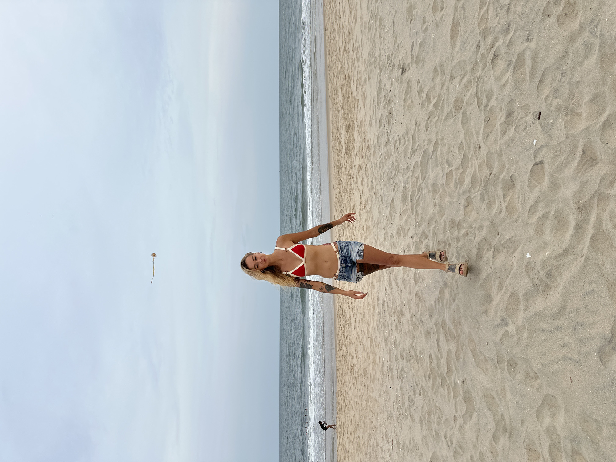 A young woman with blond hair, wearing a red bikini top and denim shorts, stands on a sandy beach. She has tattoos on her arms. The ocean is behind her. In the distance, a kite is flying. In the lower left of the image, a few people are near the water. Based on the location request, this image is likely taken in or near Phu Thuy Ward, Vietnam.