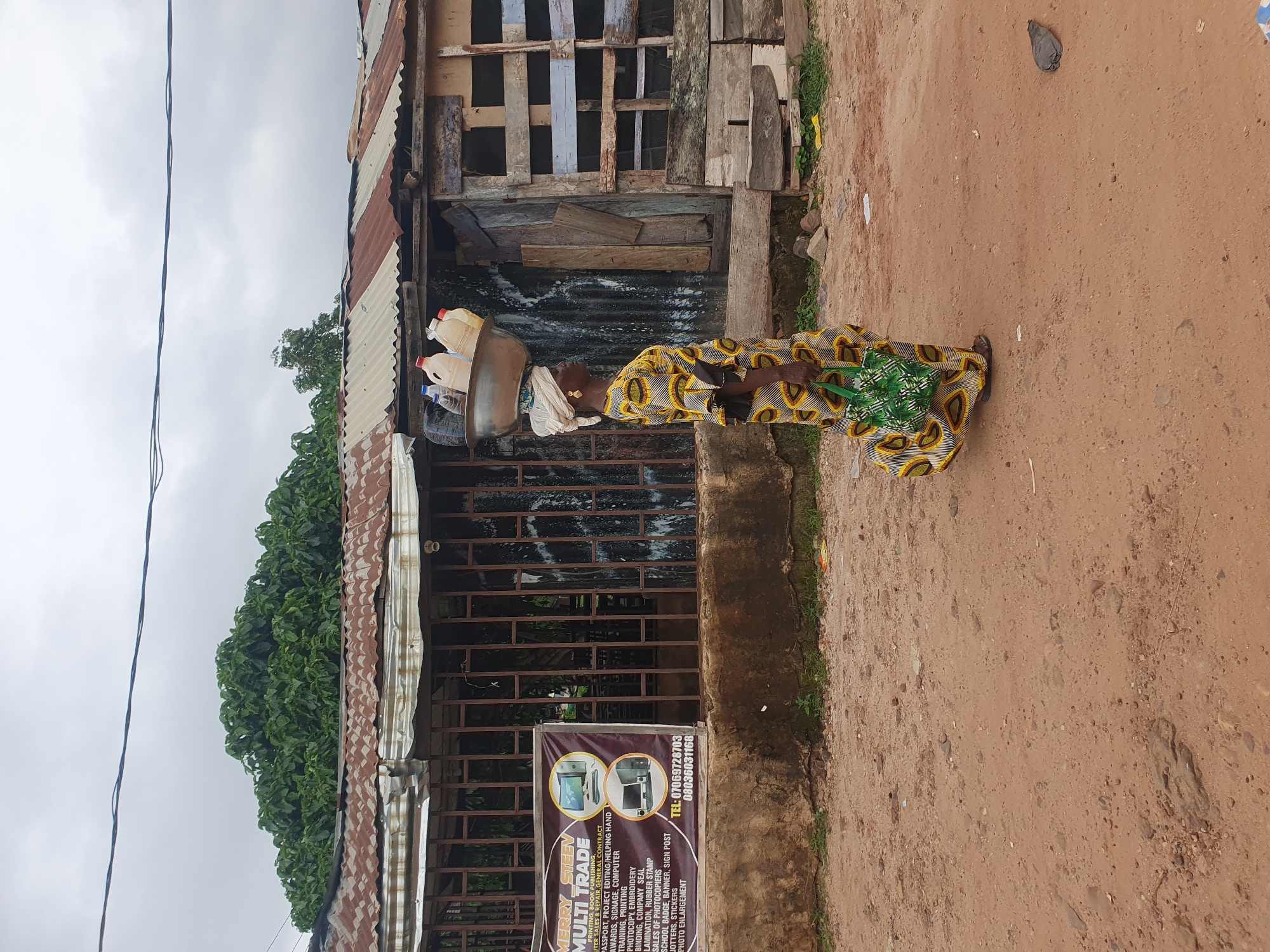 A woman is walking on a dirt road in Lokoja, Nigeria, carrying a large metal basin filled with plastic bottles on her head. She is wearing a vibrant yellow and black patterned dress and a white headscarf. A green patterned bag is slung over her left arm. The background features a corrugated metal-roofed building with various materials stacked against it, including wooden planks. A sign for "MERRY SLEEV MULTI TRADE" is visible on the left side of the building. The sky is overcast.