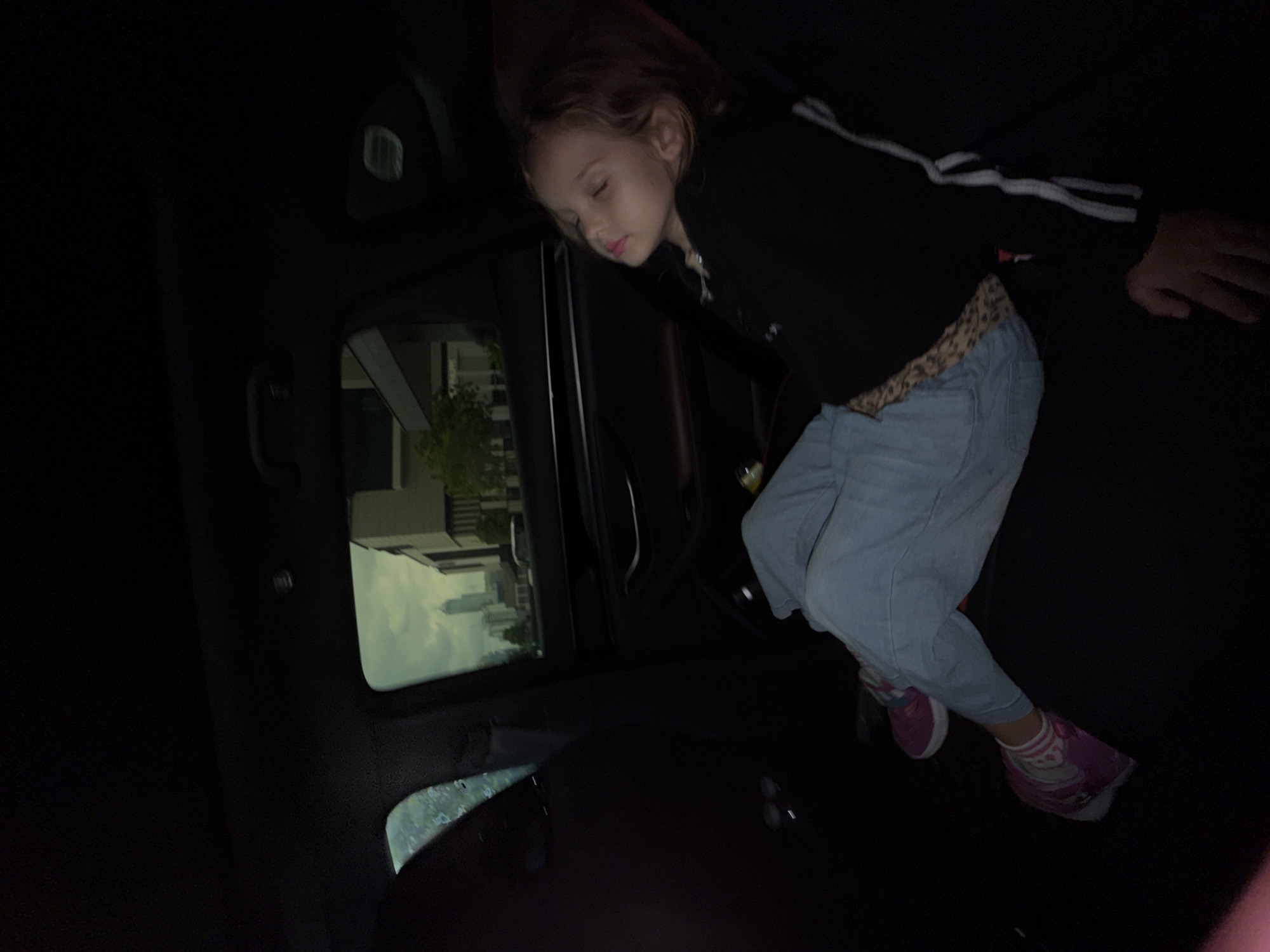 A young girl sleeps in the back seat of a car. She is wearing a black hoodie with white stripes down the sleeves, blue jeans, and pink sneakers with white socks. Her eyes are closed and her head is resting on her arm. Outside the car window, a cityscape is visible, with buildings and trees. The sky is overcast. The scene appears to be in Bangkok, Thailand.