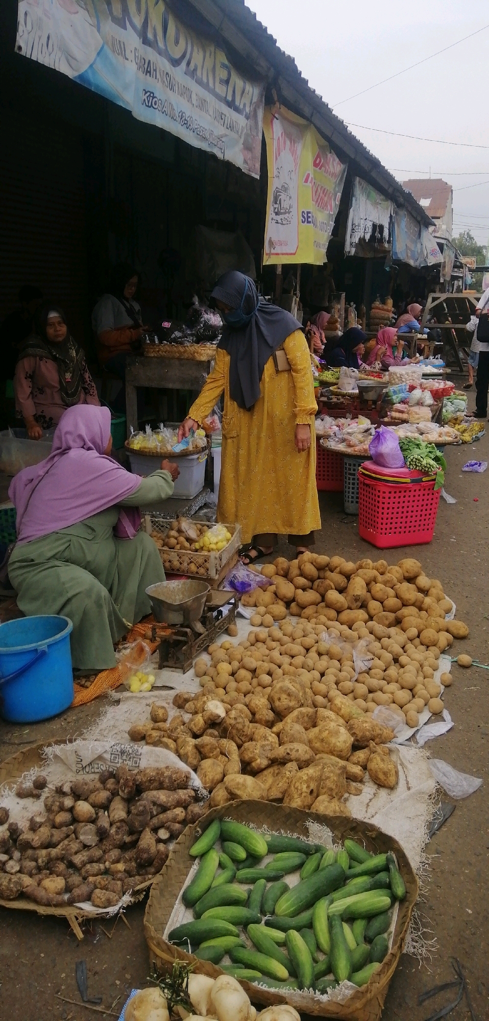 The image captures a street market scene in Ajibarang, Indonesia, with a focus on the sale of fresh produce. A woman in a yellow dress and a dark headscarf is seen interacting with another woman in a green outfit and a purple headscarf, possibly in a transaction. They are surrounded by piles of potatoes, yams, and a basket of cucumbers. Several other people, mostly women wearing hijabs, are visible in the background, either attending stalls or shopping. The market appears to be set up along a street with vendors displaying their goods on the ground and from stalls. Banners with text, some partially obscured, hang overhead, indicating the presence of businesses and potentially advertising the location as being in Ajibarang. The overall environment suggests a bustling local market atmosphere with a focus on agricultural products.