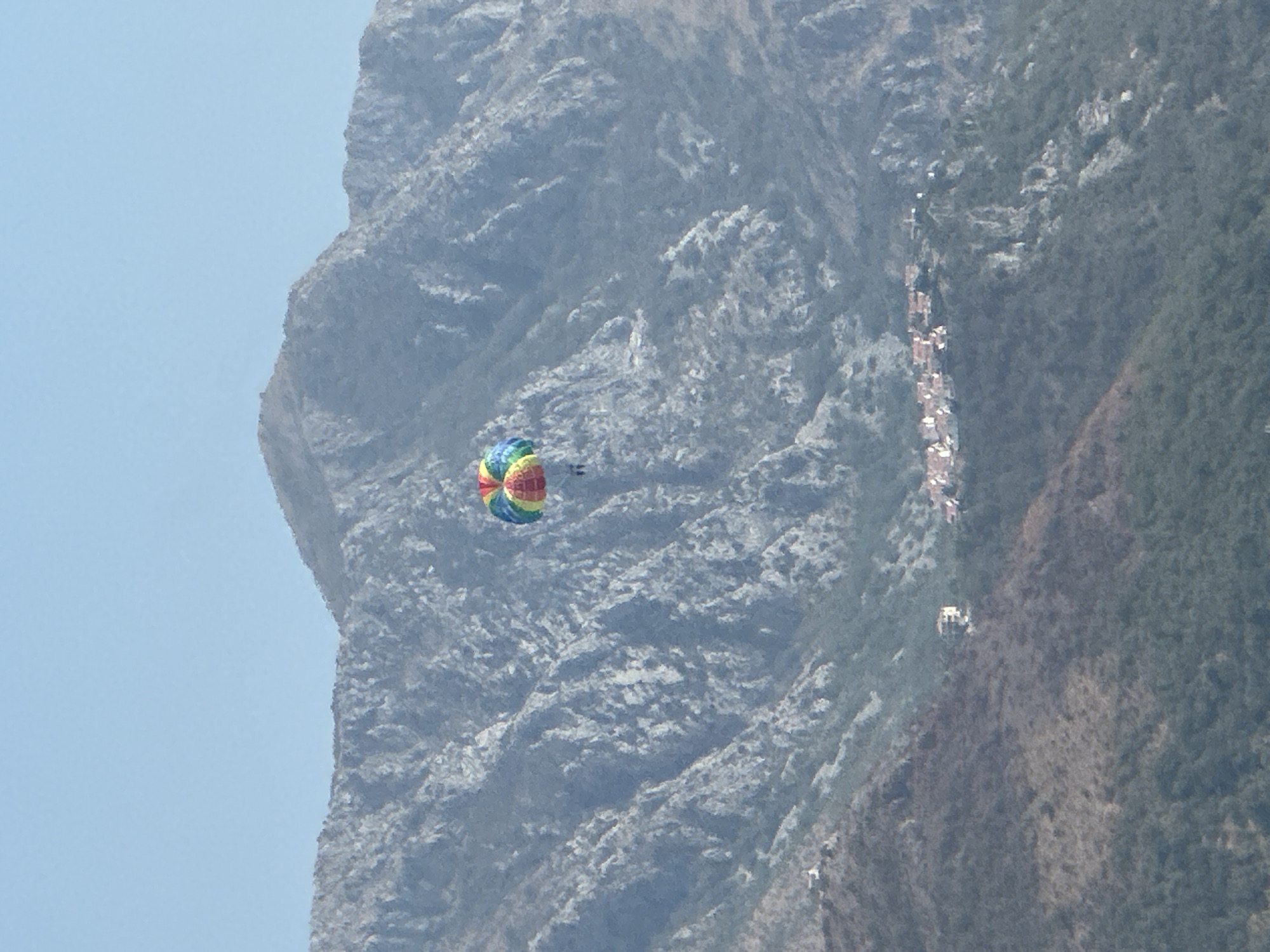 A parasailer with a colorful canopy is seen suspended against a backdrop of a steep, rocky mountain range. Below the parasailer and further down the mountain slope, a small cluster of buildings with red roofs forms a village. The foreground consists of a densely forested hillside. The sky above is clear and pale blue. The scene appears to be set in a coastal mountainous region, likely Boreti, Montenegro, given the characteristic landscape and architecture.