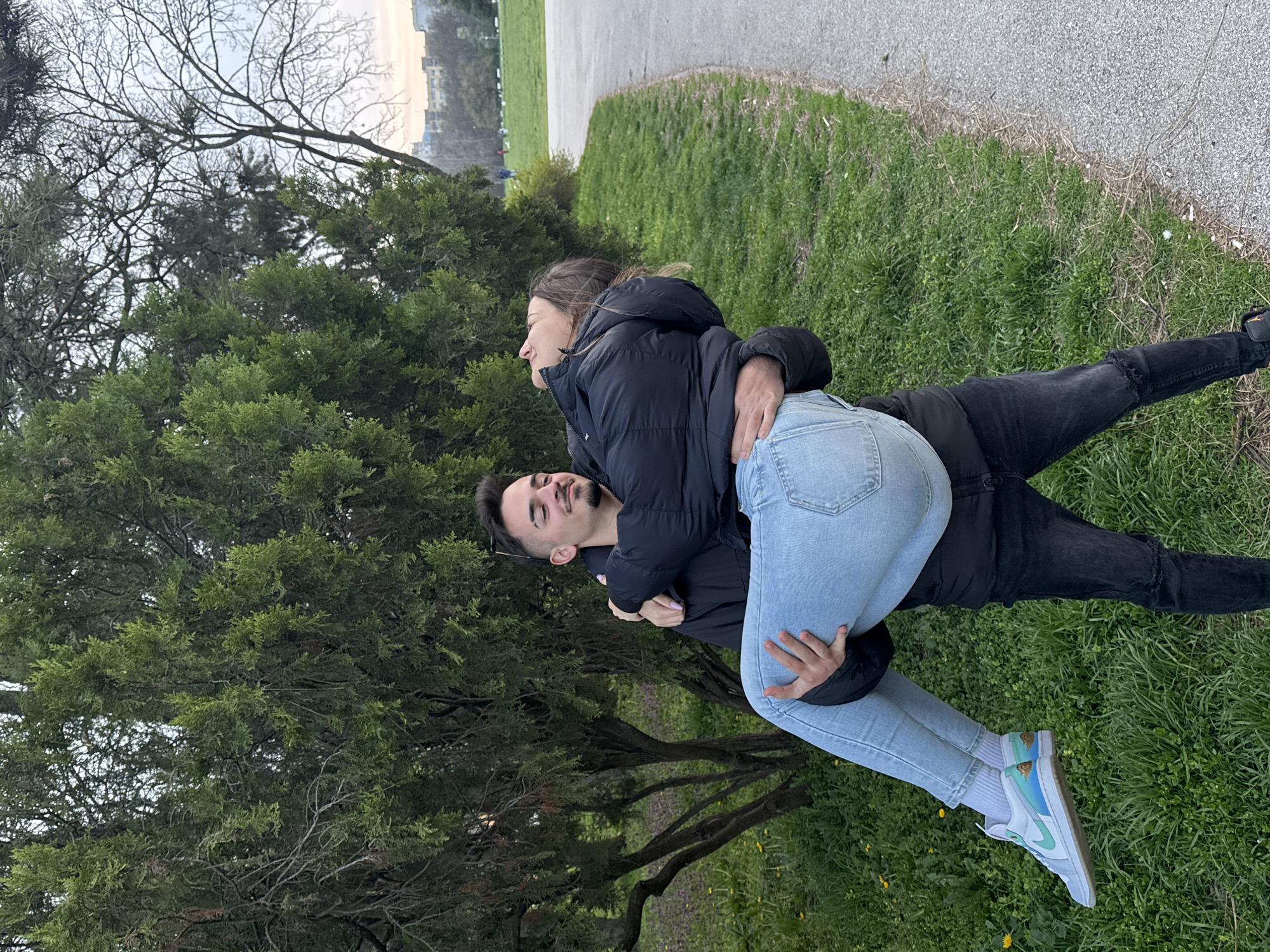 A man and a woman are embracing in Borisova Gradina Park, Sofia, Bulgaria. The man is carrying the woman in his arms, their bodies intertwined. The man is wearing a black jacket and black jeans. The woman is wearing a black jacket and light blue jeans and white and light blue sneakers. They are on a grassy area with trees in the background. A paved path is visible to the right of the grass.