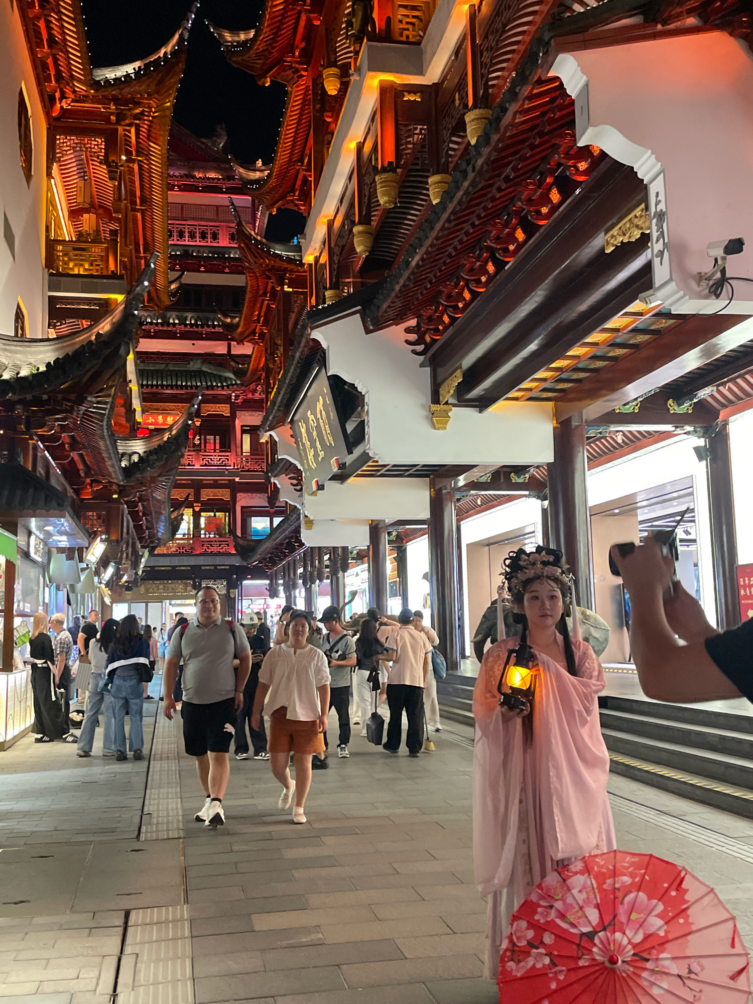 This image depicts a street scene in Shanghai, China, likely at night given the lighting. The architecture features traditional Chinese multi-tiered roofs with ornate eaves, illuminated by warm, orange lights. Buildings are constructed with dark wood beams and white walls, creating a visually striking contrast. A street, paved with grey tiles, runs through the center.
In the foreground, a woman dressed in a traditional pink garment, possibly a Hanfu, is holding an old-fashioned lantern. Her hair is styled elaborately with floral ornaments. Next to her, on the right, a red umbrella decorated with pink floral patterns rests on the ground.
Several other people are walking along the street. To the left of the woman in the pink dress, a man in a grey t-shirt and black shorts walks with a woman in a white top and orange shorts. Further down the street, a diverse group of individuals are seen in casual attire, suggesting this is a public area, possibly a market or tourist attraction.
The overall atmosphere suggests a cultural or historical district within Shanghai, characterized by traditional aesthetics combined with modern pedestrian traffic.