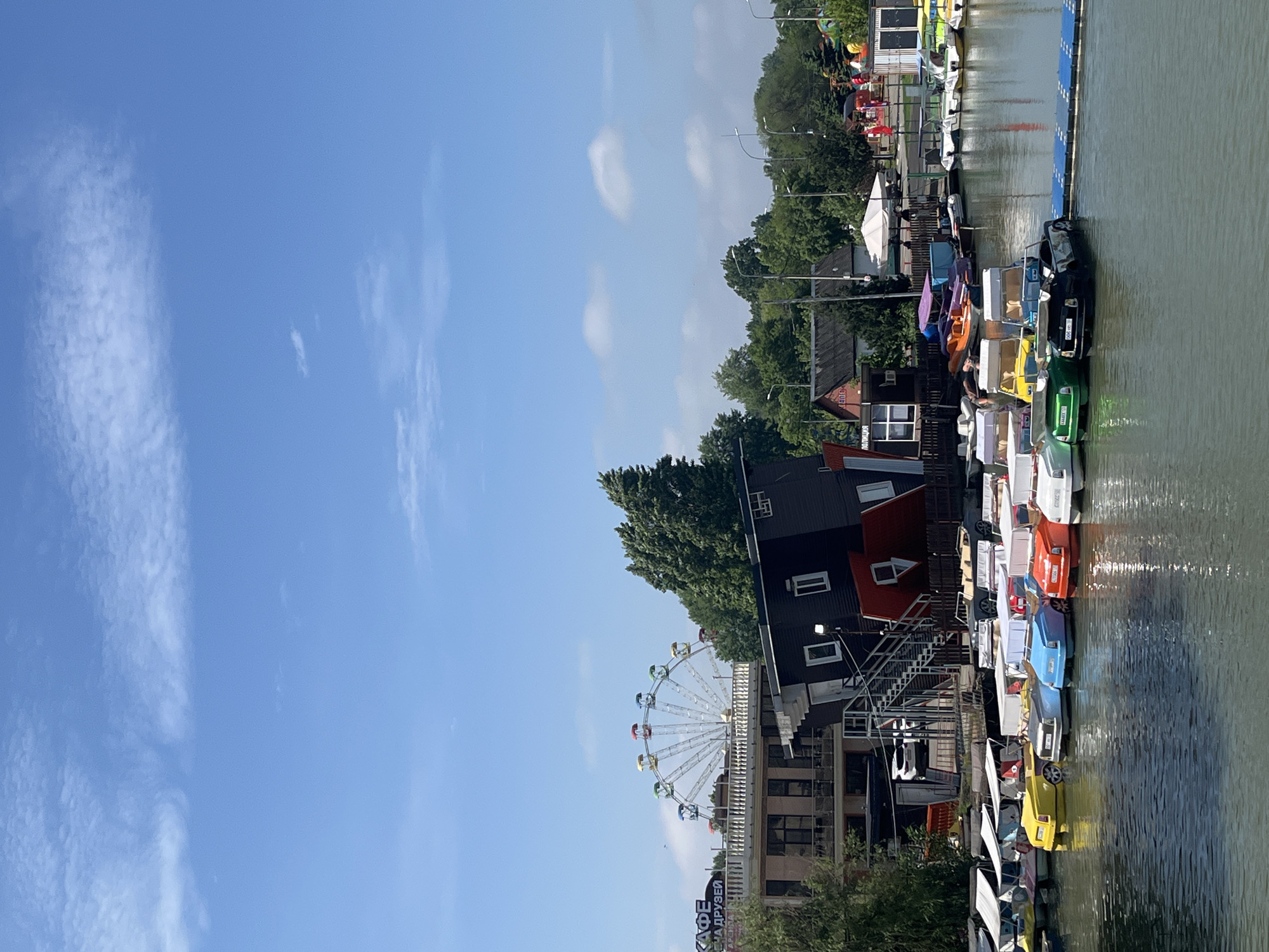 Here is a description of the image:
The image depicts a waterfront scene in Krasnodar, Russia. The focus is on a line of colorful paddle boats docked along a waterway. A pedestrian bridge extends into the frame. Behind the boats are buildings, including one with a red roof and a black exterior, with stairs leading up to it. Further back, a Ferris wheel is visible. The sky is clear and blue, with scattered clouds.