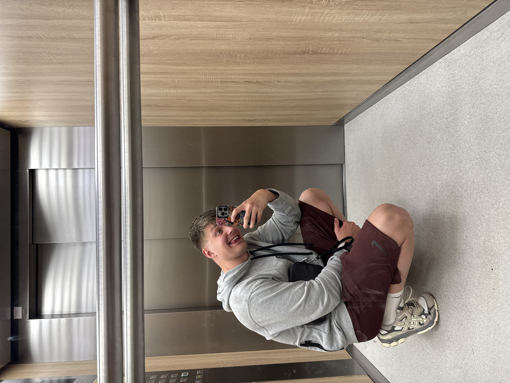 A young, Caucasian male is taking a selfie inside an elevator. He is squatting in the corner, facing the elevator door and the camera, with his phone raised in his right hand. He is wearing a grey hoodie, maroon shorts, white socks, and beige athletic shoes. A black messenger bag is slung across his body.
The elevator has wood-paneled walls and a grey, speckled floor. Stainless steel panels cover part of the interior. A control panel is visible on the elevator wall. The location cannot be determined as being Plovdiv, Bulgaria without additional information.