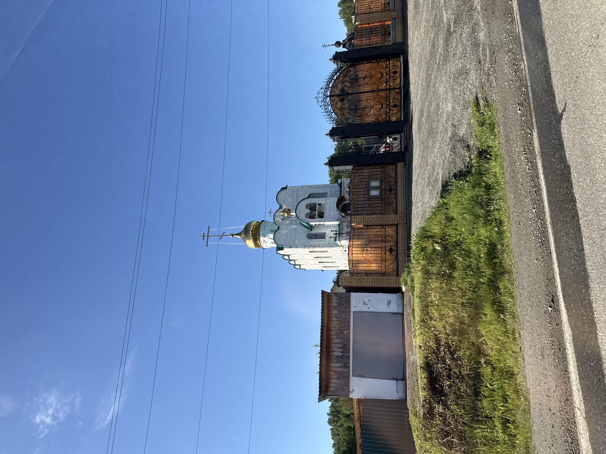 In Nizhniy Atlyan, Russia, a white Eastern Orthodox church with a golden dome and a cross stands under a clear blue sky. The church is enclosed by a brick fence with ornate golden gates and a section of metal fencing. To the left of the church, a garage with grey double doors is attached to the fence. Green grass and trees are visible behind the structures. The foreground shows a gray asphalt road.