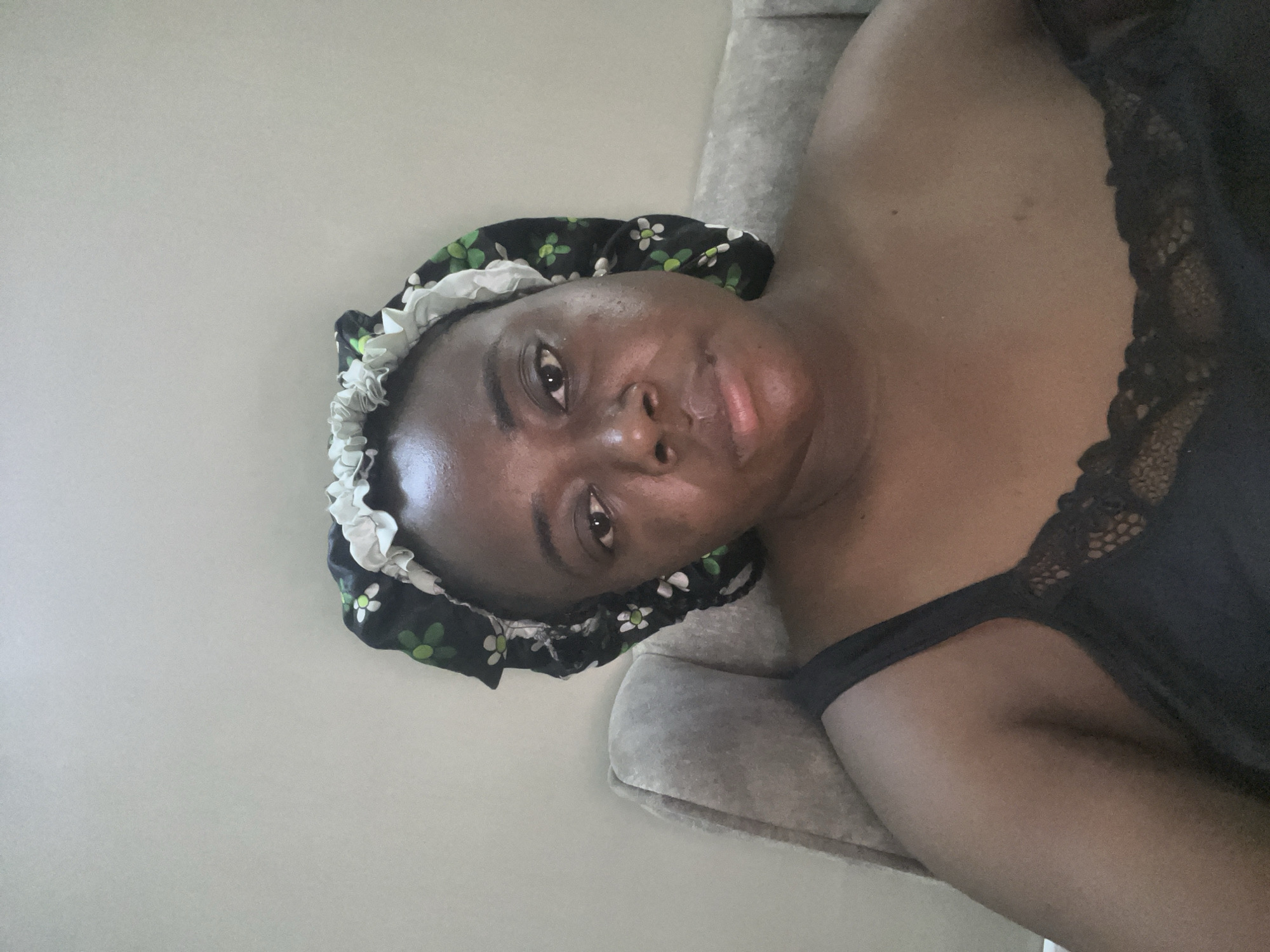 An indoor portrait features a dark-skinned individual, likely an adult female, lying down and looking directly at the camera. The image is oriented with the person on their left side.
The subject is wearing a black sleep cap or bonnet, which has a distinct pattern of small white and green flowers and a ruffled white trim along its edge. She also wears a black garment, possibly a camisole or nightgown, with intricate black lace detailing visible on the strap near her shoulder. Her expression is neutral, and her gaze is direct.
The background consists of a plain, light-colored wall or surface, behind which a textured, light gray upholstered object, possibly a sofa armrest or a bed headboard, is visible. The lighting is soft and even, suggesting an indoor setting without direct sunlight. There are no clear indicators of the time of day or external weather. No text is visible in the image. The setting appears to be a domestic environment in Abuja, Nigeria.