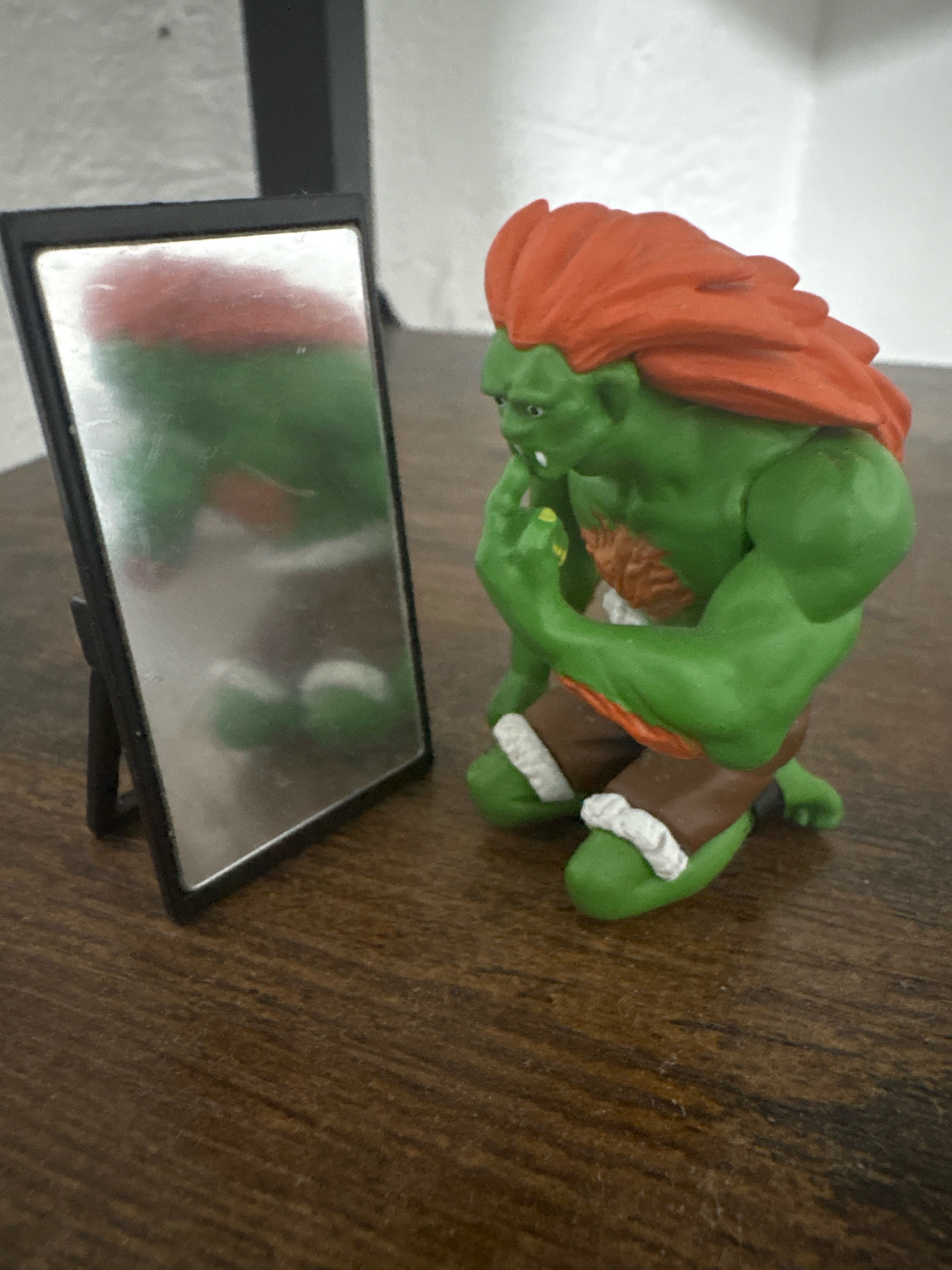 A green, muscular toy figure with bright orange hair is kneeling and looking into a small, rectangular mirror placed on a wooden surface. The figure is wearing brown shorts with white trim and has a patch of brown fur on its chest. Its left hand is raised to its face, with its index finger touching its chin. The mirror reflects the figure, showing its green skin, orange hair, and the white trim on its shorts. The background is a light-colored wall, possibly a plain textured surface, and a dark vertical object. The lighting appears to be from an overhead source, casting soft shadows. The scene is a close-up shot, focusing on the toy and its reflection. There is no information available about the location in Cuernavaca, Mexico.