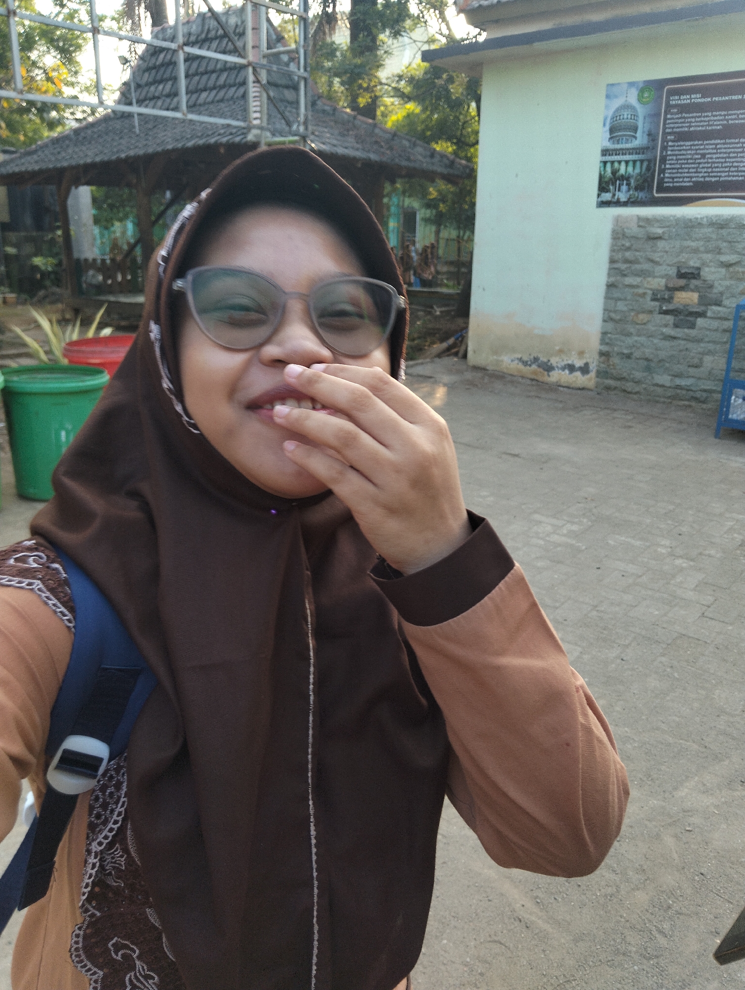 In Paciran, Indonesia, a young woman wearing glasses and a brown hijab is captured in a selfie. She is smiling, with her right hand covering her mouth, and her eyes are closed. She is wearing a brown hijab and a brown and beige patterned top, with a blue backpack visible on her left shoulder. Behind her, a traditional Indonesian gazebo with a dark roof and metal framework is visible, along with some trees. To the right of the gazebo, a light-colored building with a stone lower section stands. The ground is paved with a rough, grey surface, possibly asphalt or concrete, with some scattered debris. The scene appears to be outdoors, with natural light indicating daytime.