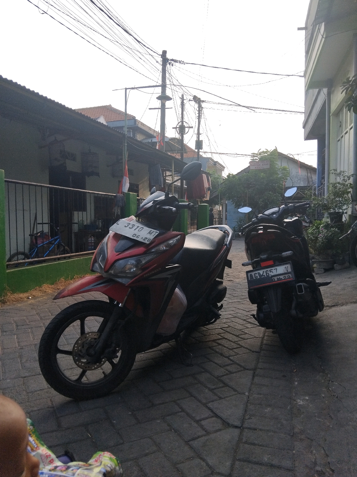 The image depicts an outdoor scene on a paved pathway in Kepuh Kiriman, Indonesia, likely a residential area. Two motorcycles are parked on the pathway. The motorcycle in the foreground is red and white, with a license plate displaying "L 3310 MT 06-28". The second motorcycle, in the background, is darker in color, possibly black or dark gray, with a license plate reading "AG 4657 V".
Adjacent to the pathway is a green fence, behind which is a building with a corrugated roof. A bicycle with blue handlebars is visible behind the fence. The sky is bright and overcast, suggesting daytime. Overhead, a complex network of electrical wires crisscrosses the sky, supported by utility poles. To the right of the motorcycles, part of a building with white columns and windows is visible. Vegetation, including trees and potted plants, is present in the scene. In the lower left corner, the blurry foreground shows a glimpse of a baby's head and a colorful object, possibly a stroller or a baby carrier. A sign is visible on a building in the background which reads "PELAN-PELAN BANYAK ANAK KECIL", translating to "Slowly, many small children".