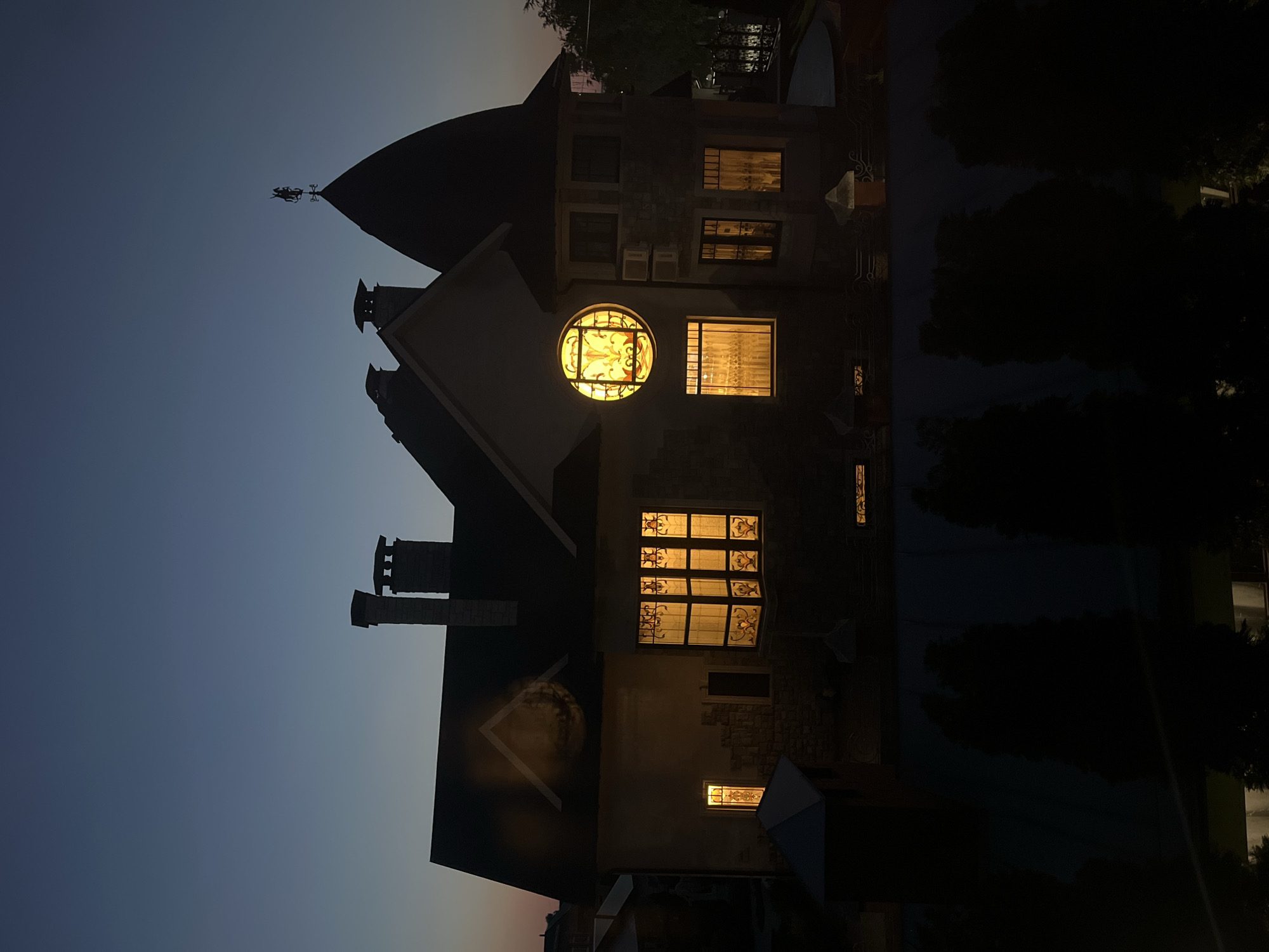 This is a nighttime exterior, eye-level, medium shot of a multi-story stone and stucco house with a dark roof, likely in Balakovo, Russia. The sky is a deep gradient of twilight blue. The house features several prominent architectural elements: two brick chimneys, a conical turret topped with a weather vane depicting a knight on horseback, and a distinctive round stained-glass window with yellow and orange hues. Light emanates from multiple windows, casting a warm glow on the facade. The windows are adorned with ornate patterns, particularly the large circular one. In the foreground, silhouetted trees obscure the lower portion of the house and the surrounding landscape, which appears to be a lawn or garden. The overall impression is one of a grand, possibly historic, residence at dusk.