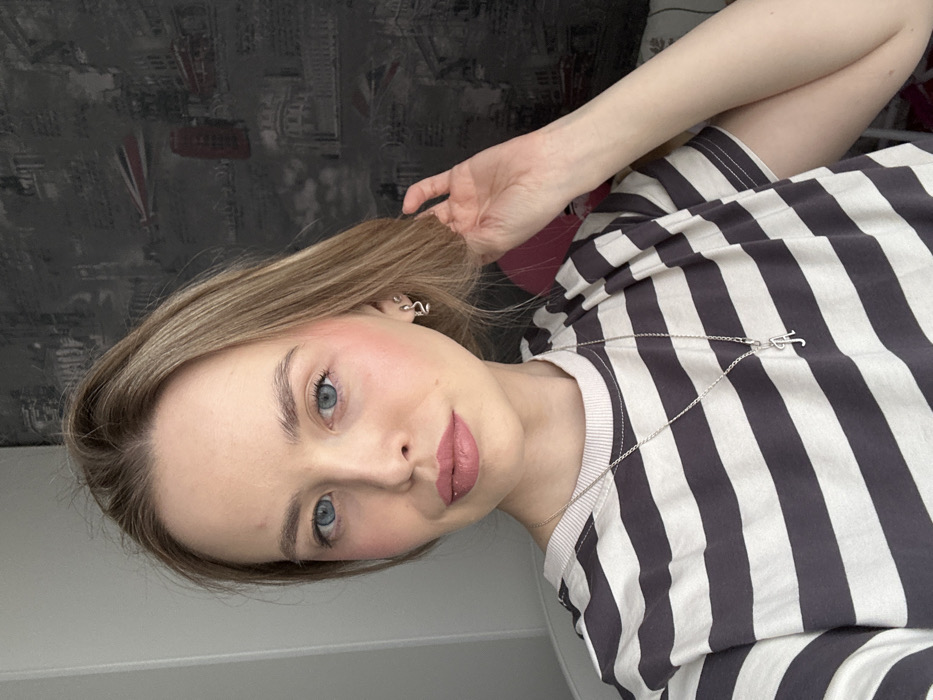 The image depicts a close-up, chest-up view of a young adult female, likely indoors. She has fair skin, blue eyes, light brown or blonde hair, and is wearing light makeup including pink blush and lipstick. Her hair is parted slightly to the left (viewer's right) and falls loosely around her shoulders. She wears a short-sleeved t-shirt with horizontal dark gray and light gray stripes. A silver-colored chain necklace with a cursive "A" pendant is visible on her chest. In her right ear (viewer's left), she wears a small, silver-colored earring, possibly stylized as a snake or 'S' shape. Her left hand (viewer's right) is raised, with fingers gently touching the hair near her right ear. She is looking directly at the camera with a neutral to slight smile. The background on the left consists of a plain light-colored wall. To the right, a darker wallpaper is visible, featuring a grayscale pattern of architectural landmarks, including a red double-decker bus, suggesting a London cityscape theme. The setting is an interior space; no visual cues are present to confirm the specific city as Chaikovsky, Russia.