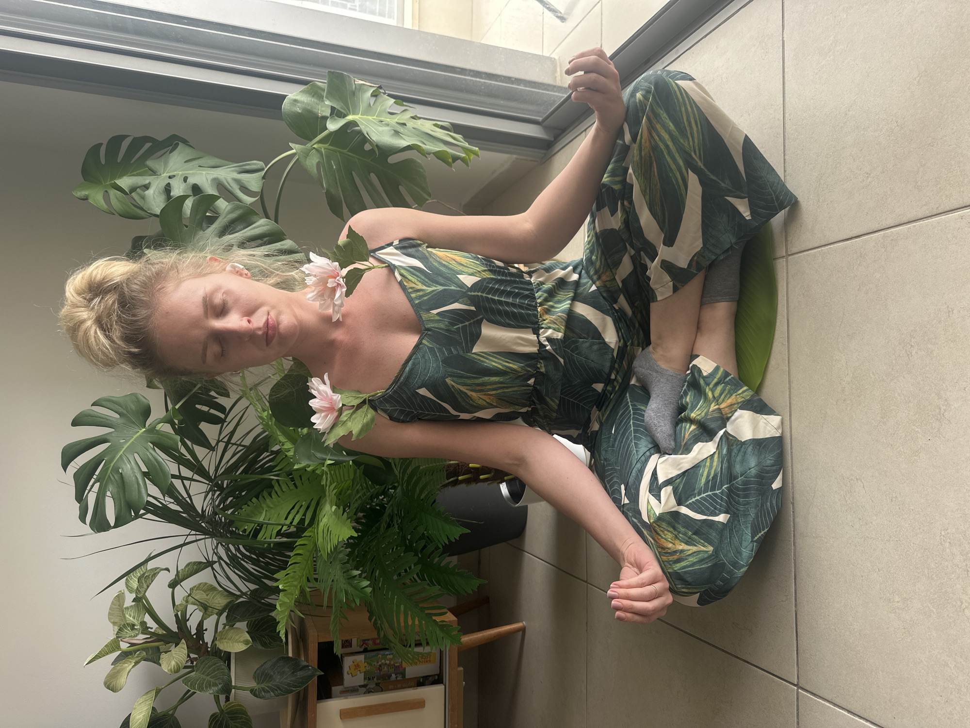 A woman with blonde hair pulled back into a bun is seated on a tiled floor in a yoga position. She is wearing a green and white leaf-patterned jumpsuit and grey socks. Pink flowers are attached to the straps of her jumpsuit. She is positioned in front of a large potted Monstera plant and other greenery. A sliding glass door and window are behind the plants. The location is likely indoors, possibly in Petah Tikva, Israel, but this cannot be confirmed from the image alone.